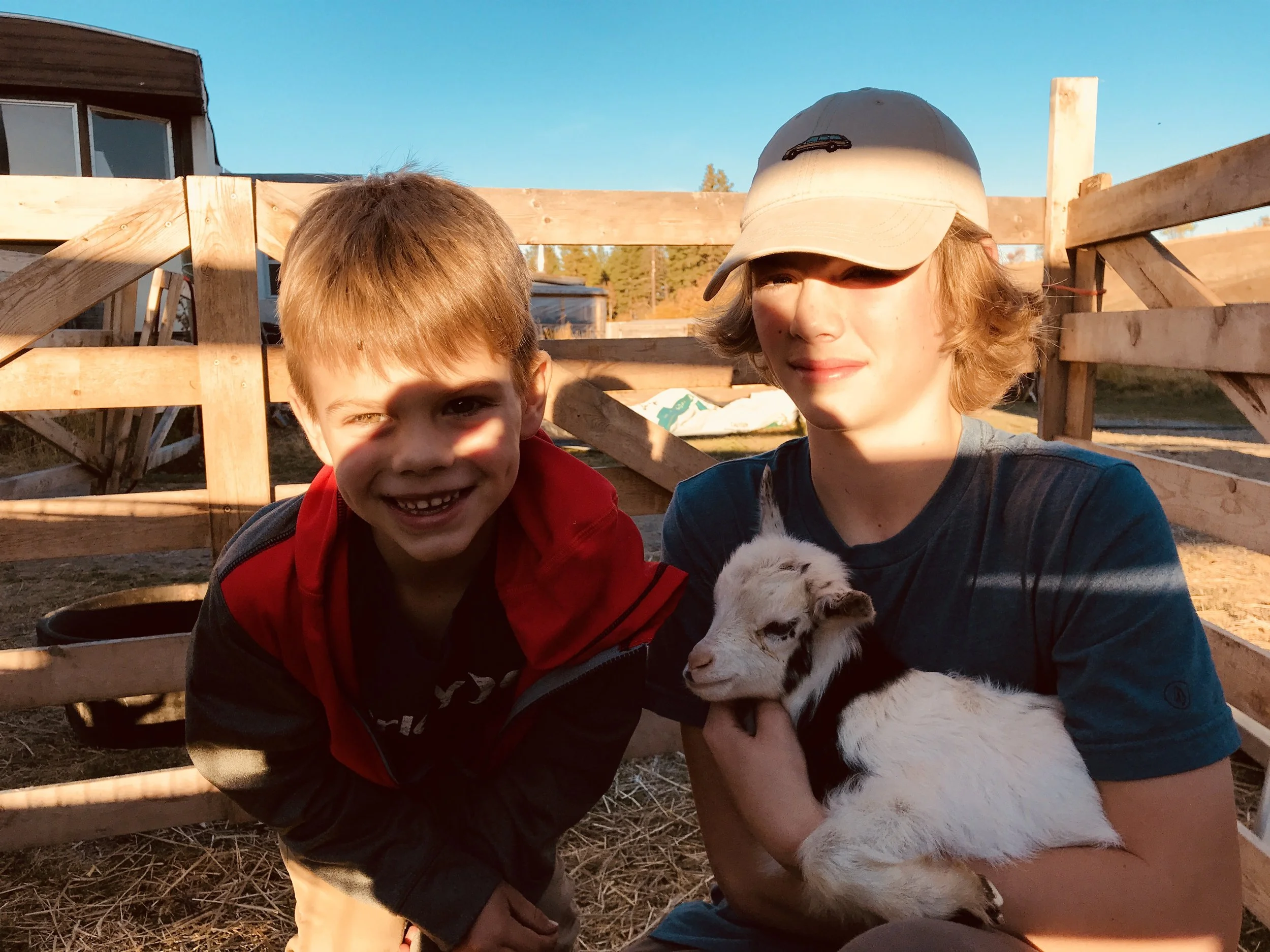 Two boys smiling and holding a puppy in front of a wooden fence during sunset or late afternoon.