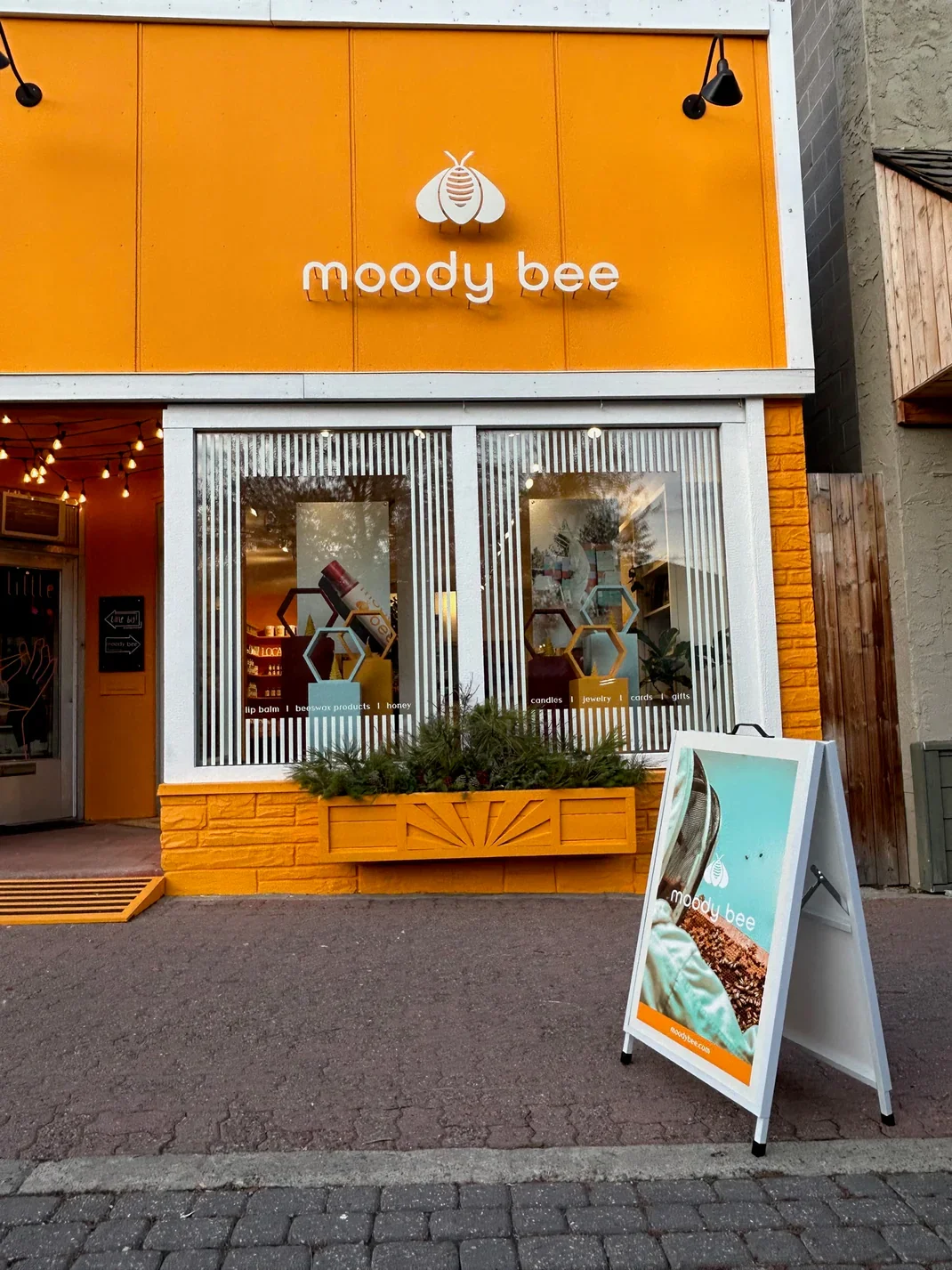 The exterior of a shop named 'moody bee' with a yellow and orange facade, a sign with a bee logo, and a sidewalk signboard showing the shop's name and products like candles, jewelry, and gifts.