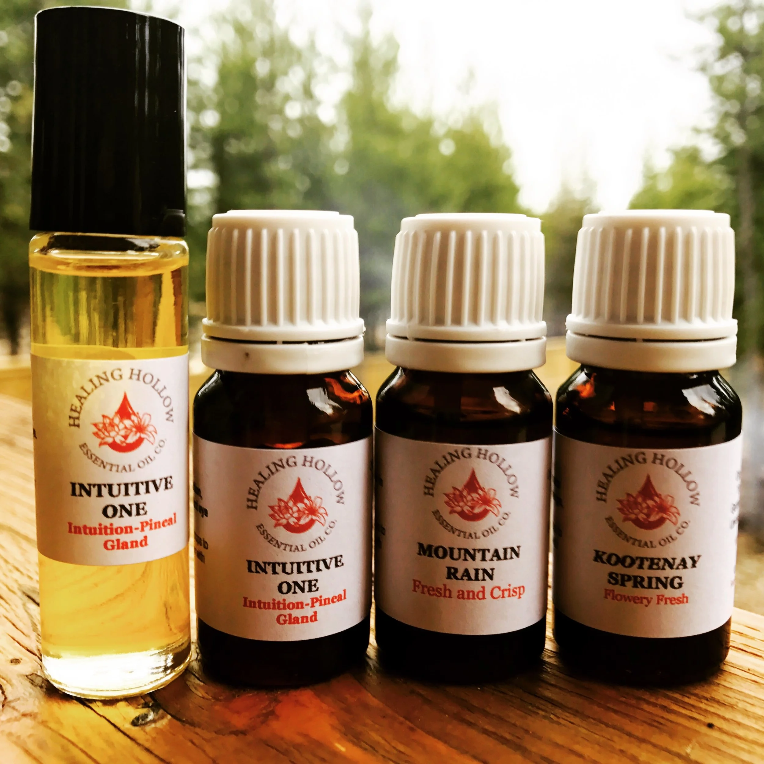 Four essential oil bottles with white labels and black caps placed on a wooden surface, with a blurred background of trees and sky.