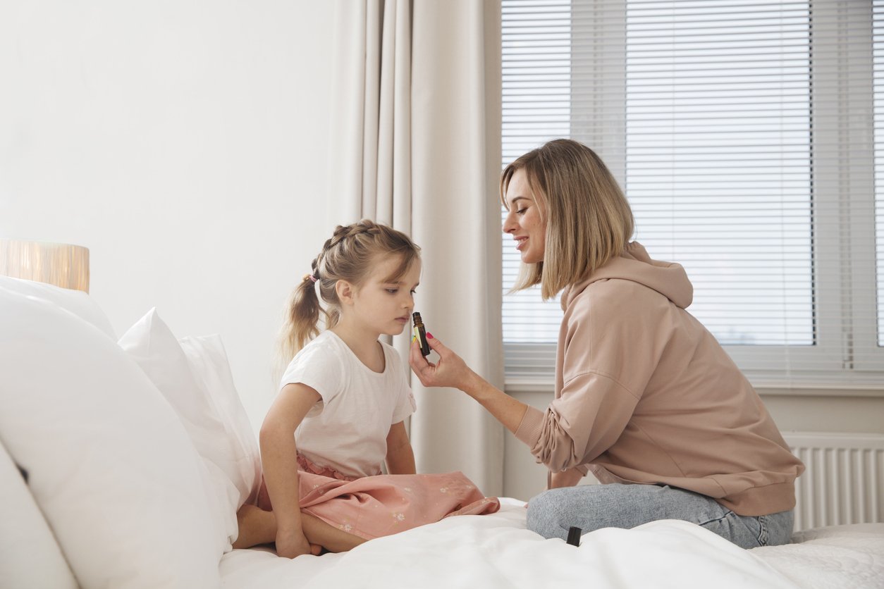 A woman giving a young girl medicine on a bed in a well-lit bedroom.