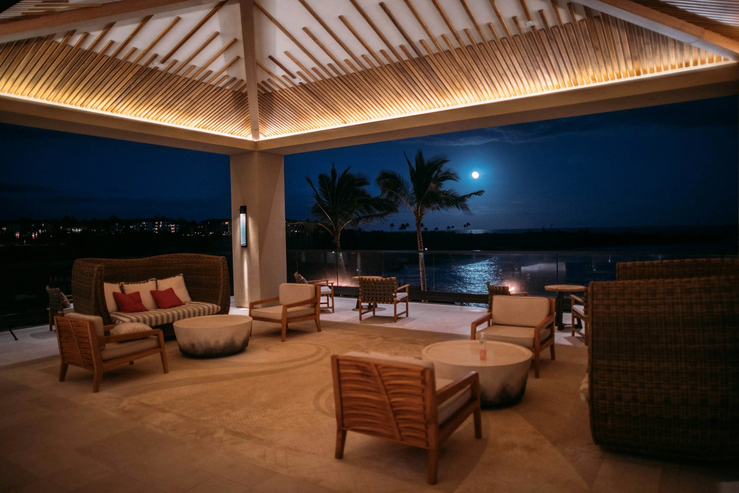 Outdoor seating area at night with wicker and cushioned furniture, trees, water, and a full moon in the dark sky.