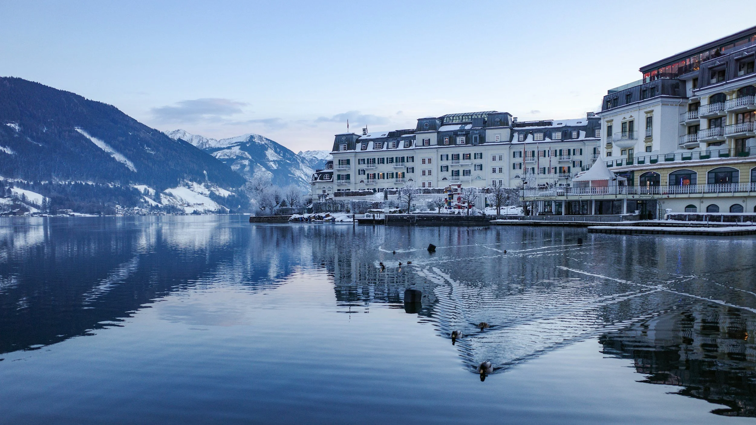 A snowy lakeside scene with large, elegant buildings along the shore, mountains in the background, and ducks swimming in the water.
