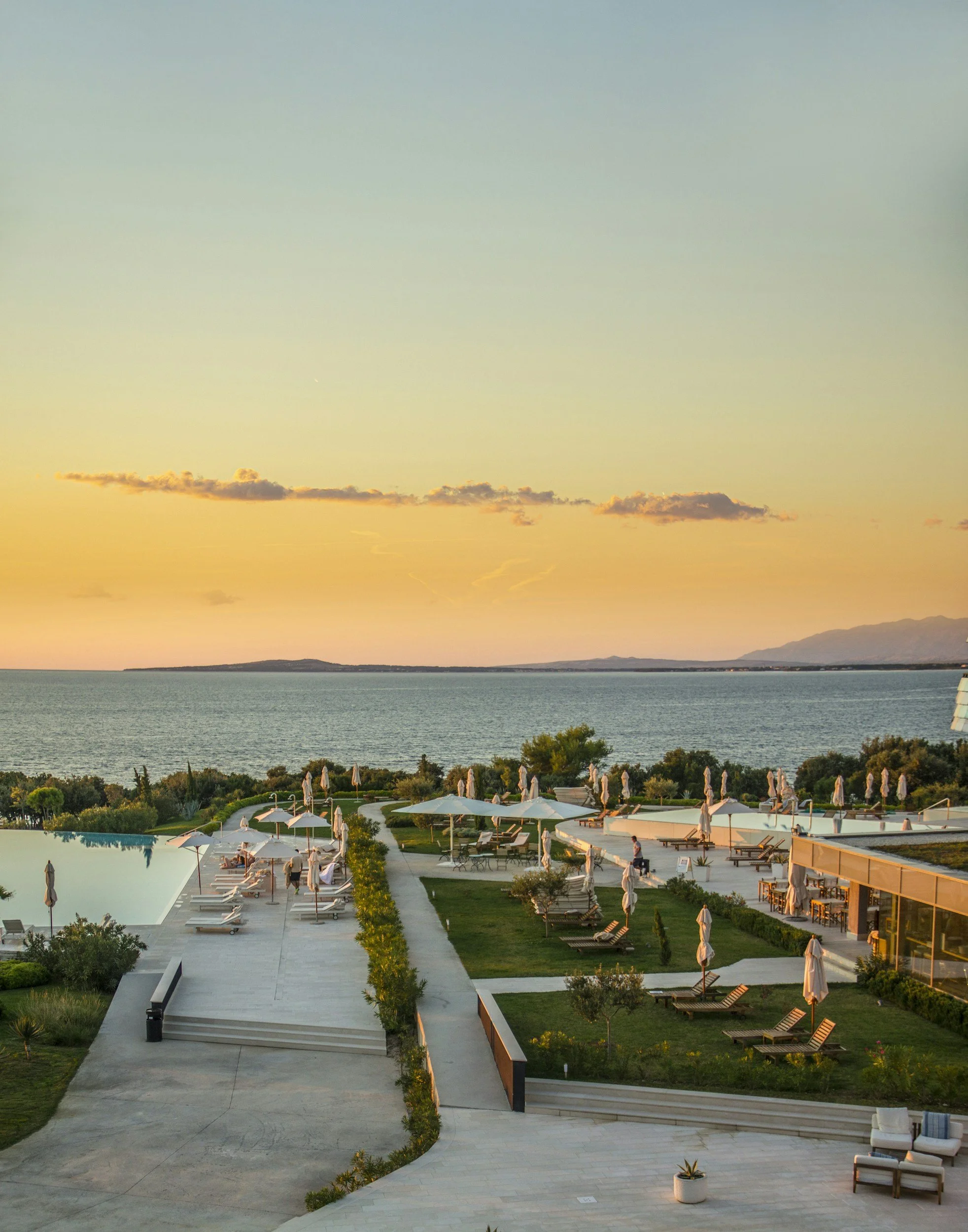 A scenic view of a coastal area during sunset, with a large poolside area in the foreground, lounge chairs and umbrellas, lush greenery, and a calm body of water with mountains in the background.