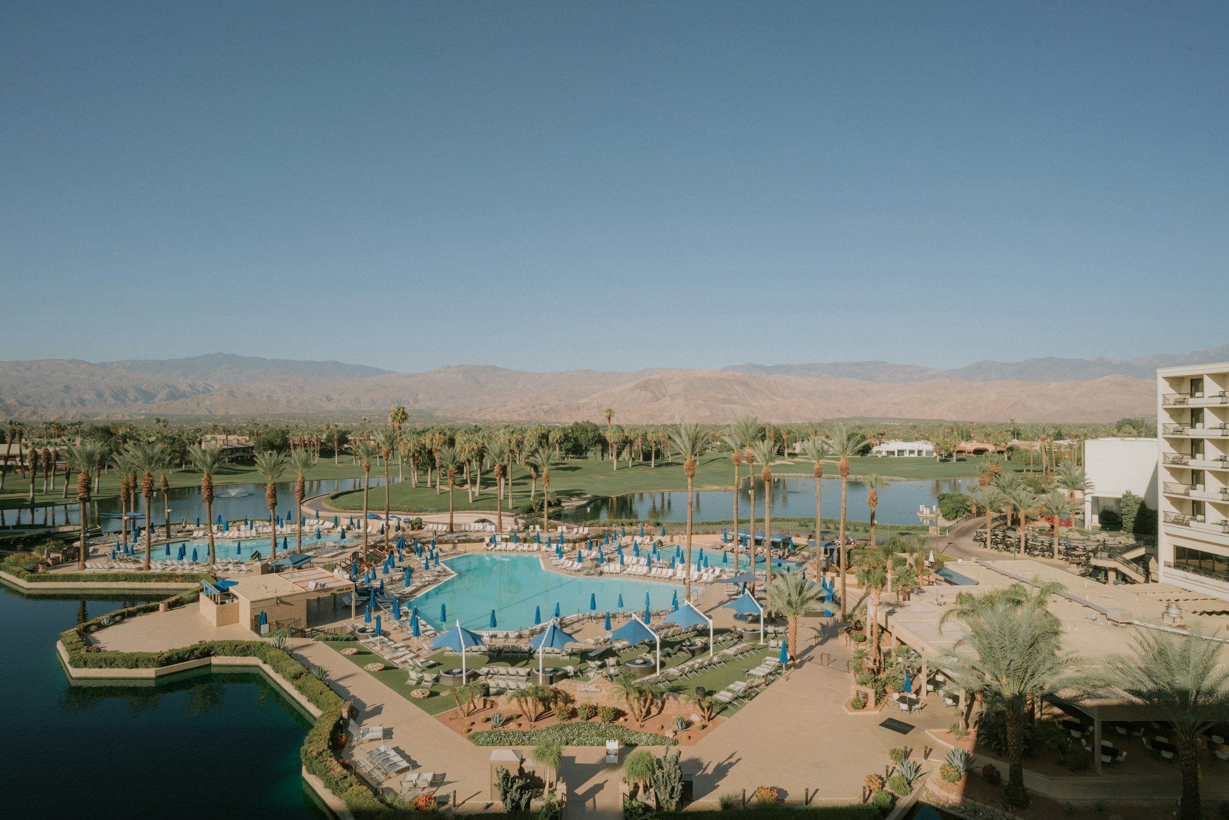 A resort pool area with numerous blue umbrellas, lounge chairs, palm trees, and a small lagoon, with mountains in the background under a clear blue sky.