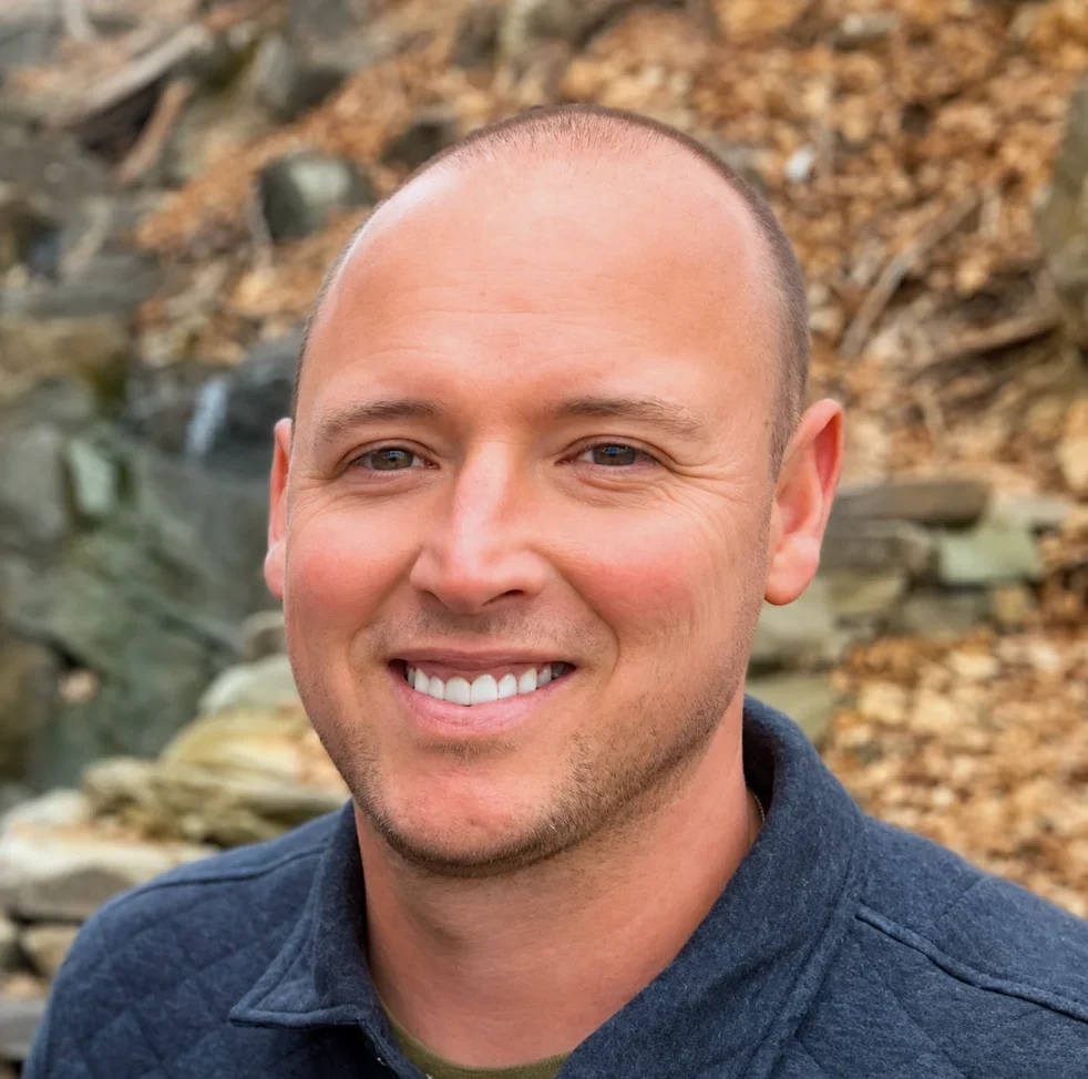 A smiling man with a shaved head and blue eyes outdoors, with a background of rocks and autumn leaves.