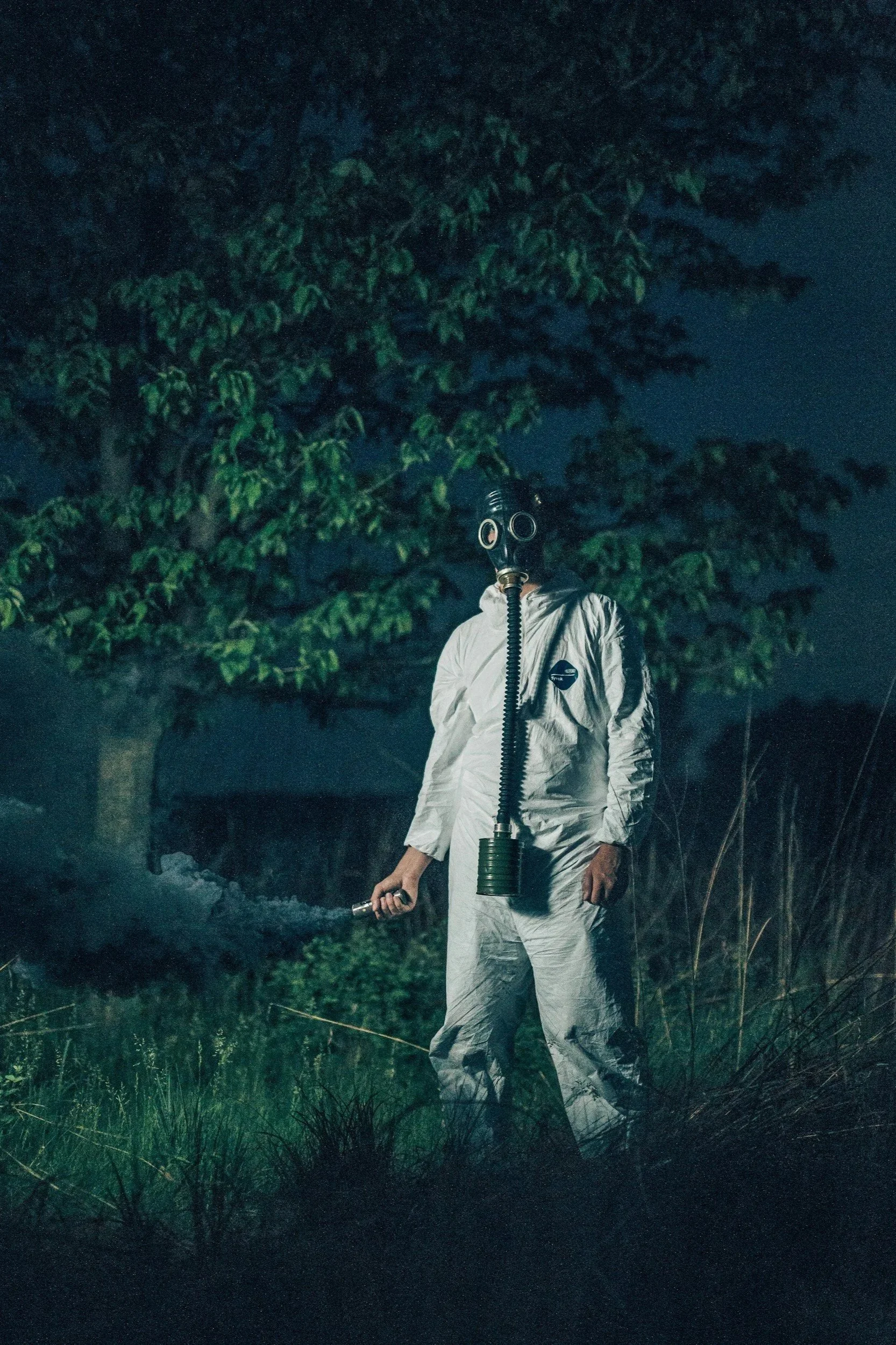 Person in protective white suit and gas mask holding a spray gun, standing outdoors at night surrounded by trees and grass, with a foggy or smoky atmosphere.