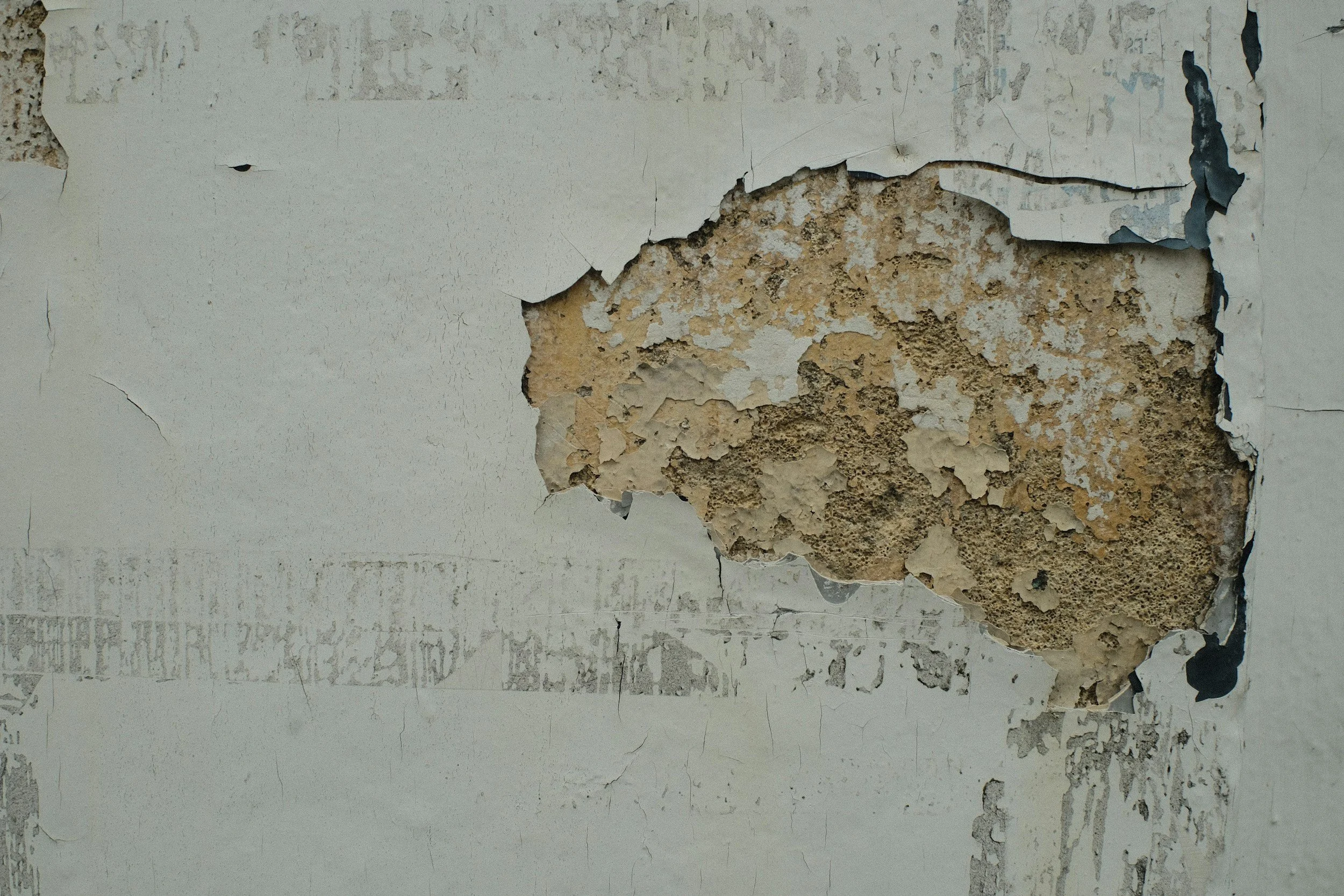 Close-up of a wall with peeling and chipped white paint revealing the underlying rough, textured brown and beige surface.