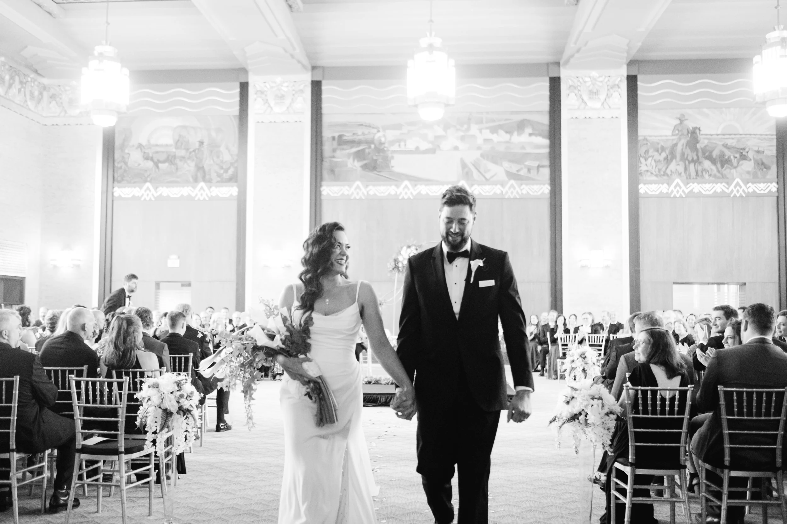 A bride and groom walking hand in hand at their wedding reception, smiling. The bride is in a white wedding dress and holding a bouquet. The groom is in a tuxedo. The reception is indoors with many guests seated at tables decorated with flowers, and large murals on the walls. Bright hanging lights illuminate the scene.