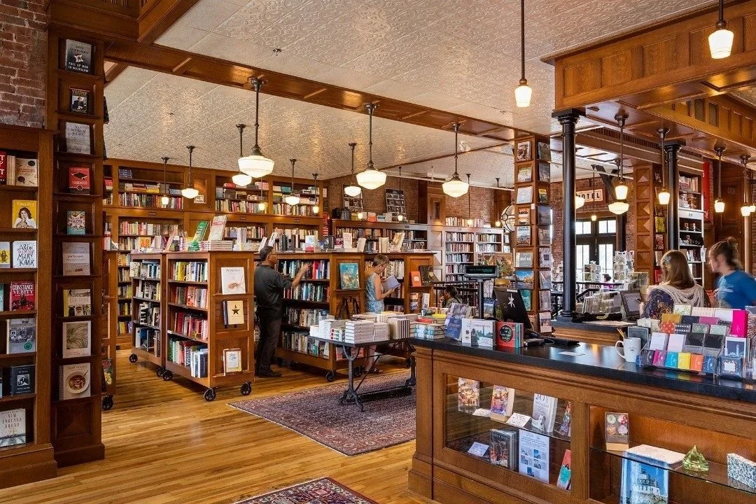 Interior of a bookstore with wooden shelves filled with books, several customers browsing, and warm lighting.