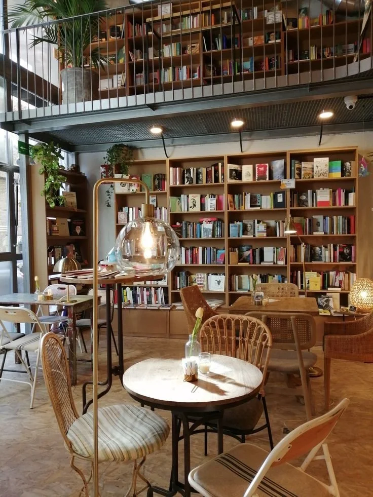 Cozy bookstore or cafe with wooden bookshelves filled with books, round tables, and various chairs, illuminated by warm lighting and natural light from large windows.