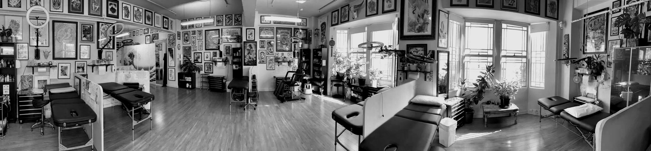 A tattoo studio with multiple tattoo chairs and tables, framed artwork on the walls, shelves with supplies, and large windows allowing natural light.