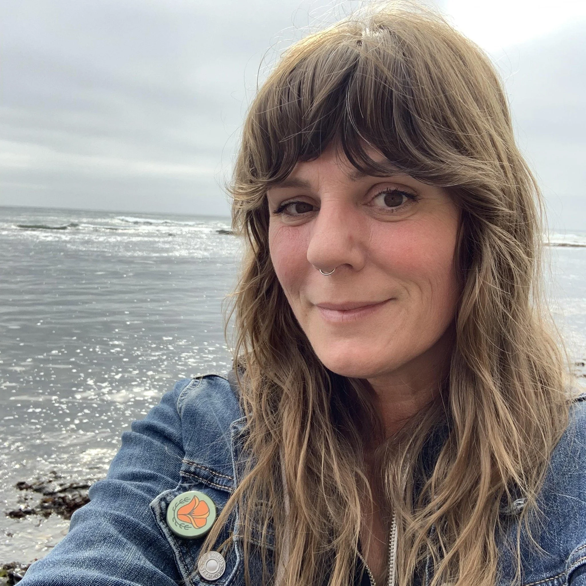 A woman with light brown hair and brown eyes at the beach with overcast skies, wearing a denim jacket with a circular flower pin on the lapel.