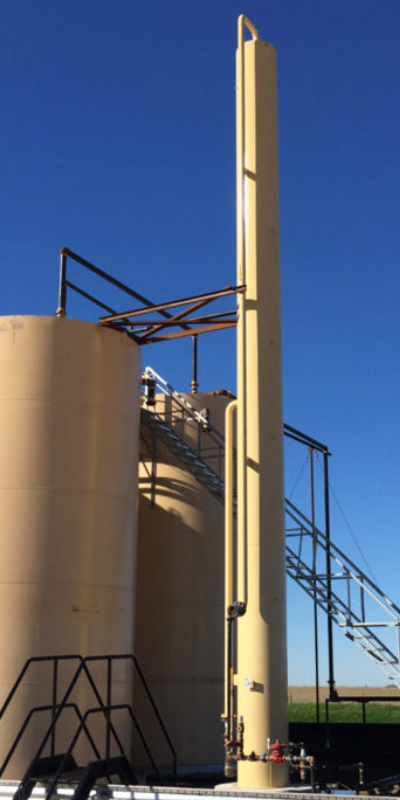 Industrial storage tanks with a tall beige exhaust stack and metal walkways against a clear blue sky.