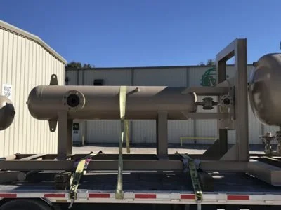 Large cylindrical industrial equipment secured on a flatbed truck outside a metal building under a clear blue sky.