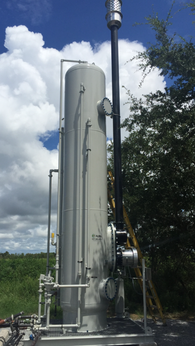 Industrial processing equipment with pipes and a tall vertical tank outdoors, against a partly cloudy sky and some trees.