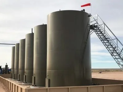 Multiple large black storage tanks outdoors with a staircase and a red flag on top, under a partly cloudy sky.
