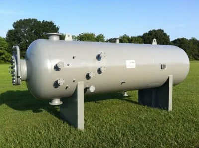 Large horizontal industrial storage tank situated outdoors on grass, with trees and a blue sky in the background.