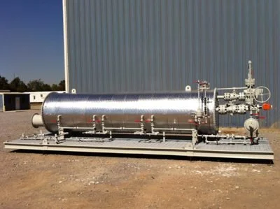 Large horizontal stainless steel industrial tank with pipes and valves outside near a corrugated metal building.
