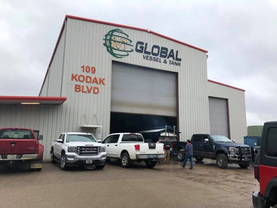 Large industrial building with a sign that reads 'Global Vessel & Tank' and the address '109 Kodak Blvd'. Several trucks and vehicles parked outside, with people working near an opened garage door.