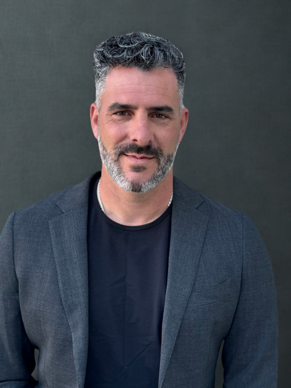Portrait of a middle-aged man with short, curly gray hair and a beard, wearing a dark blazer over a black shirt, standing against a plain dark background.