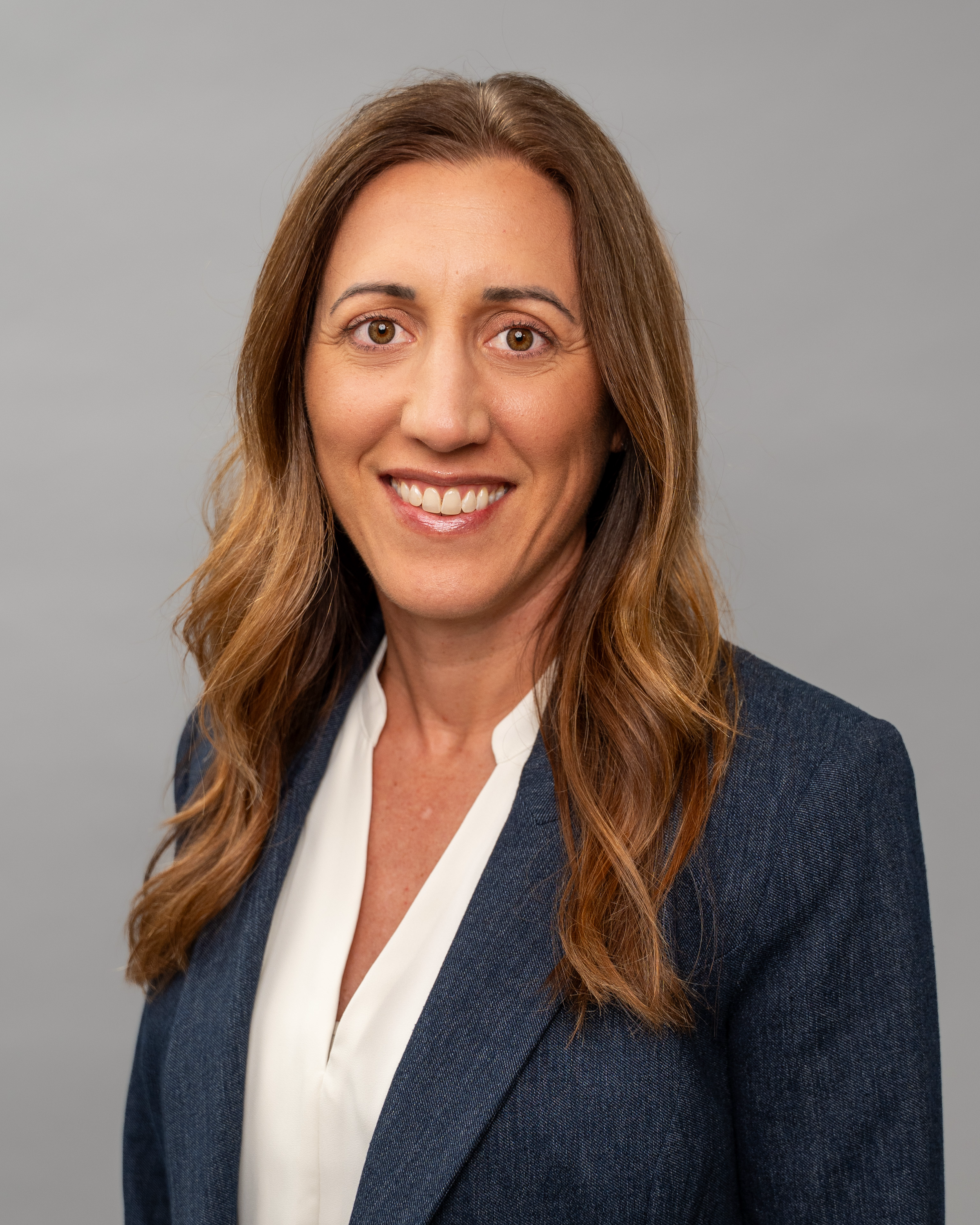 A woman with long brown hair smiling, wearing a navy blazer and white blouse, against a plain gray background.