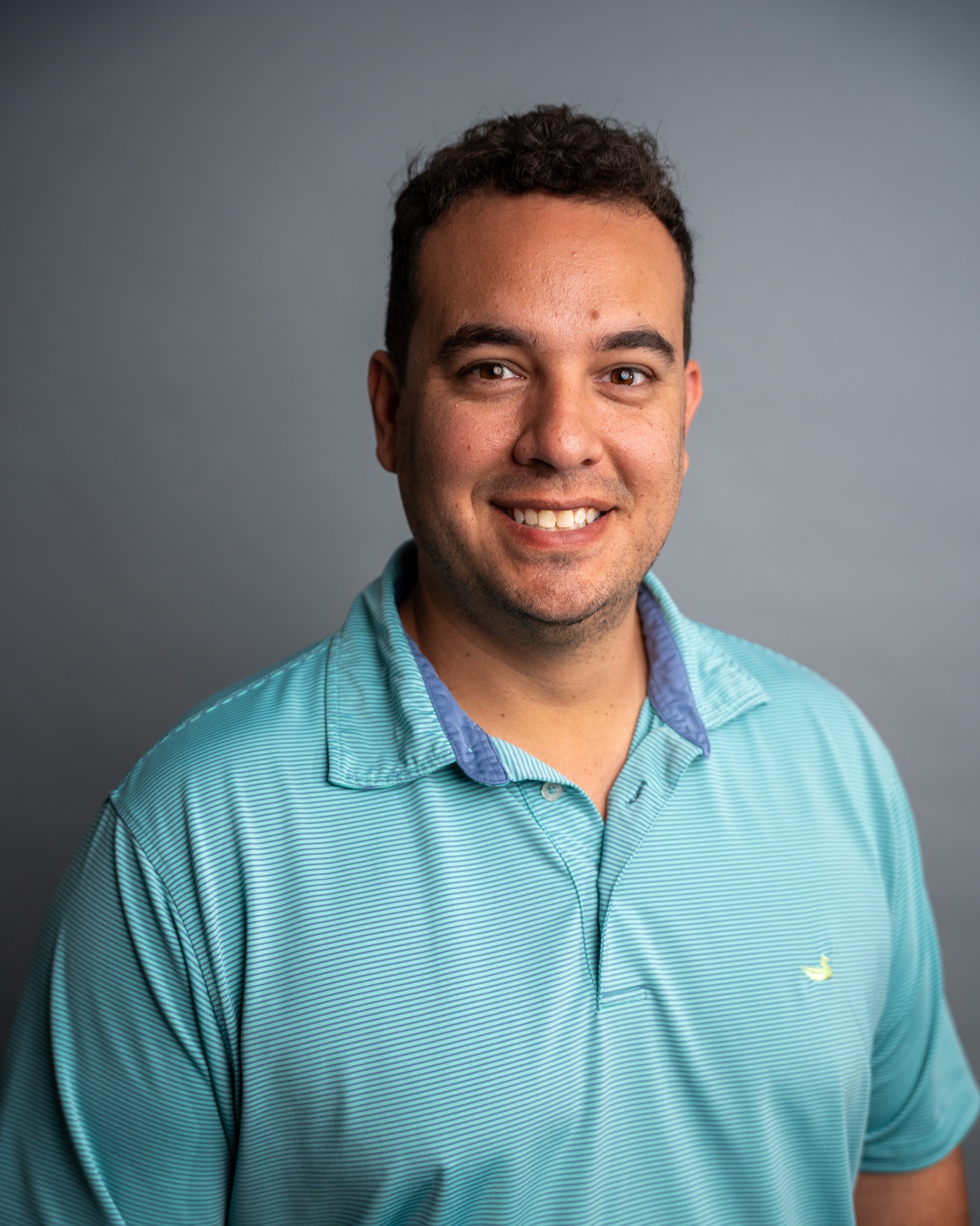 Close-up of a smiling man wearing a teal button-up shirt with thin white stripes and a small yellow logo of a duck on the chest, against a plain gray background.