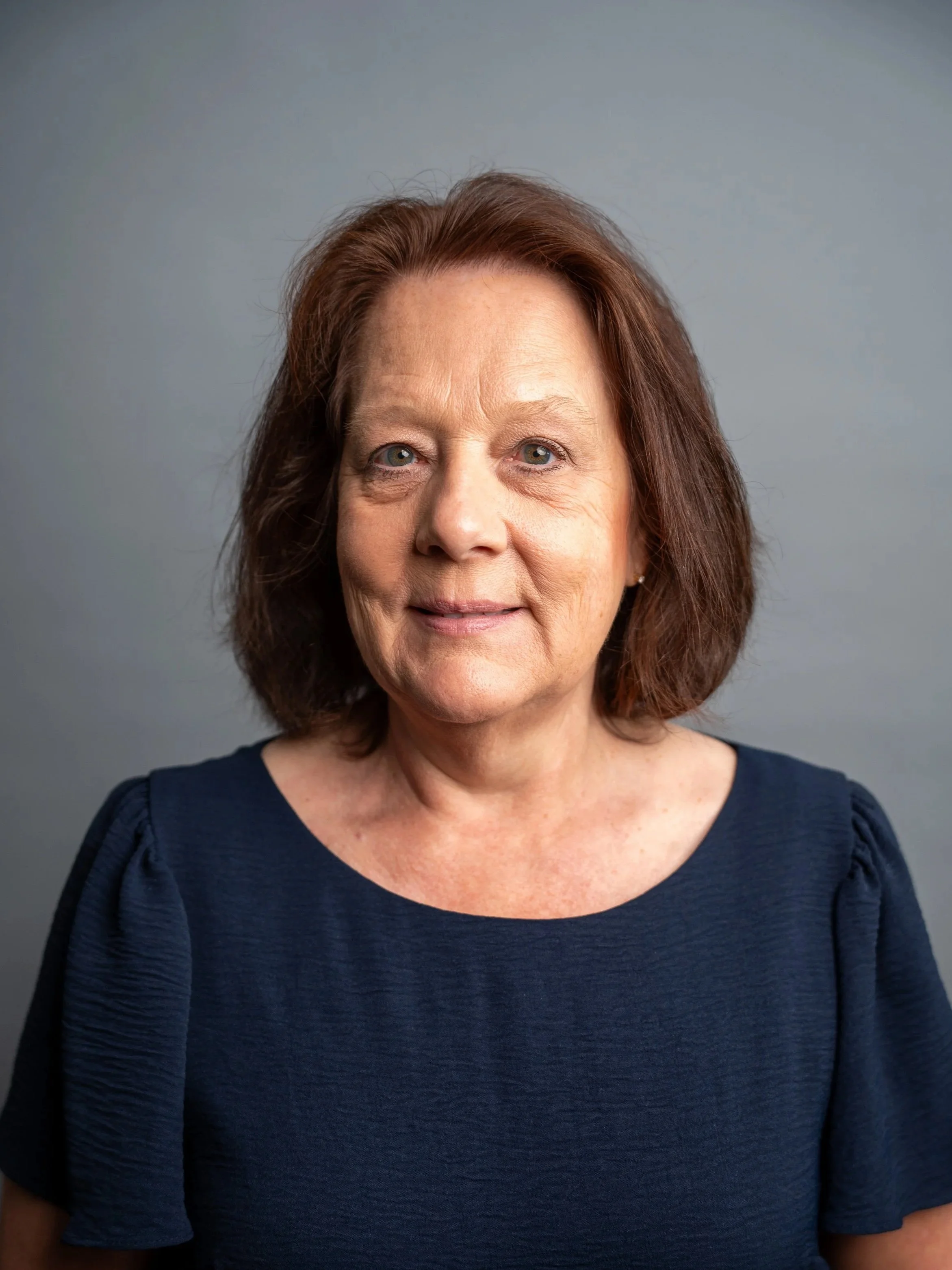 Close-up portrait of a middle-aged woman with light skin, shoulder-length brown hair, and blue eyes, wearing a navy blue top, standing against a plain gray background.