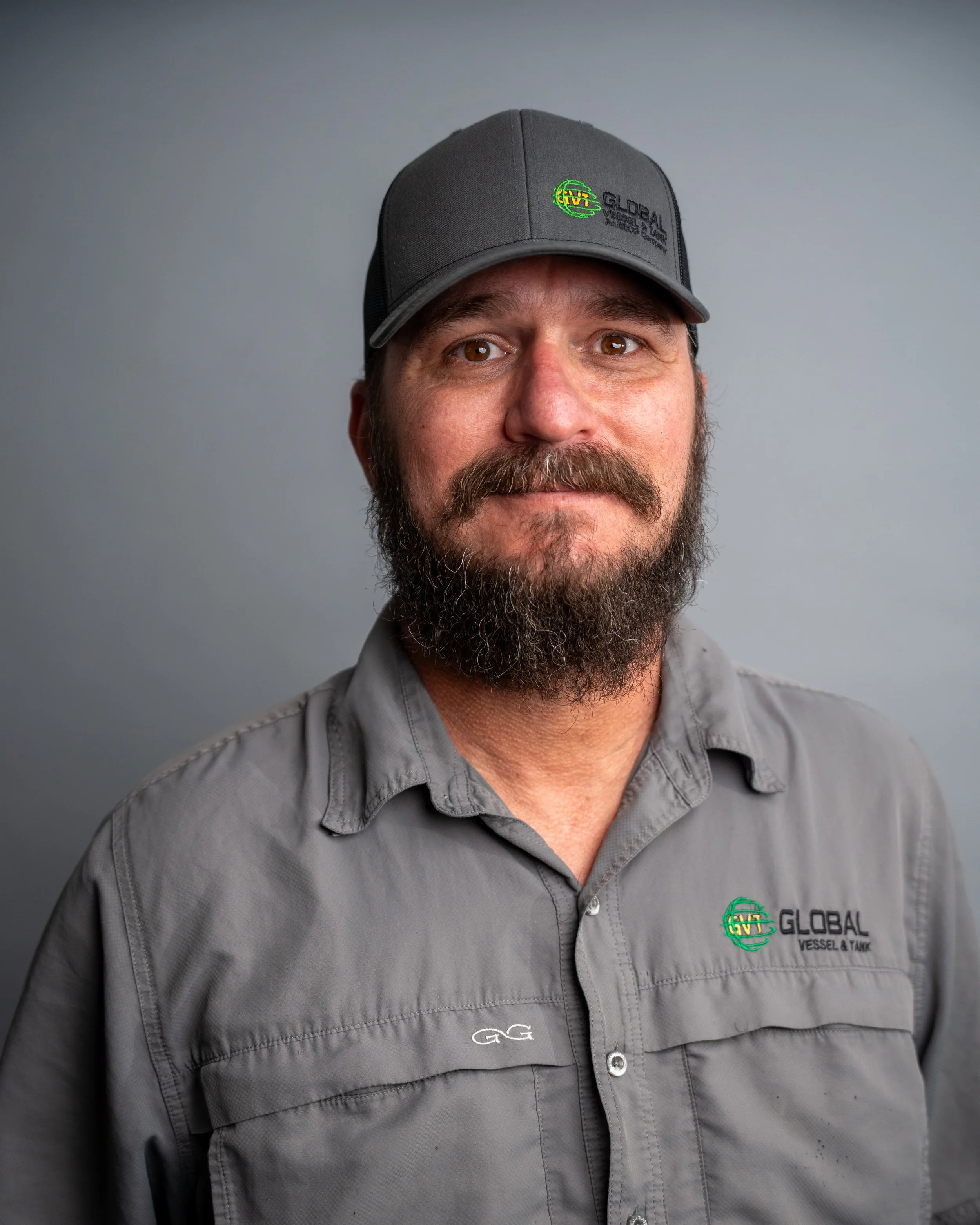 Close-up of a man wearing a gray cap with a logo and a gray shirt with a logo, against a plain gray background.
