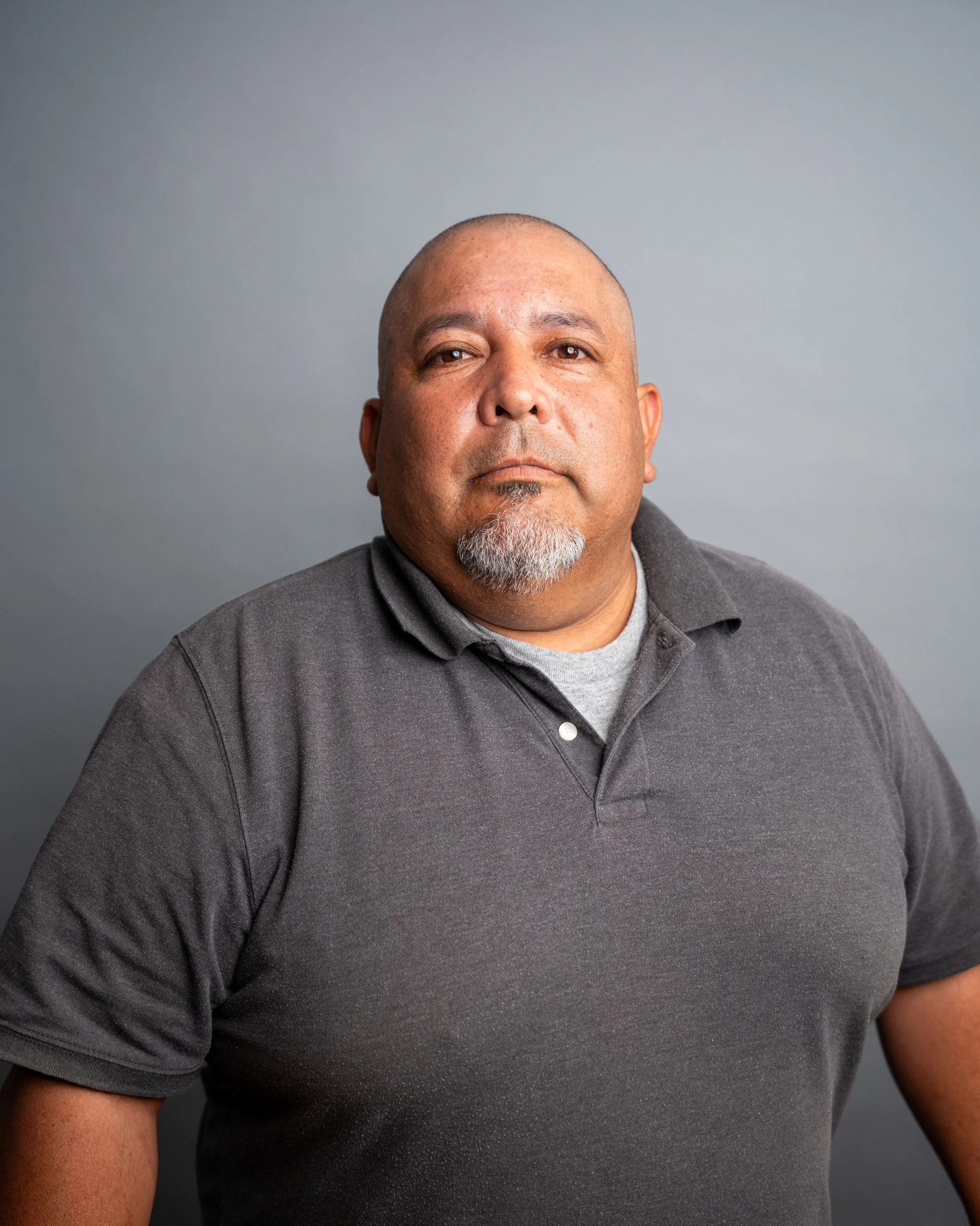 A middle-aged man with a shaved head and gray goatee wearing a dark gray polo shirt, looking directly at the camera against a plain gray background.