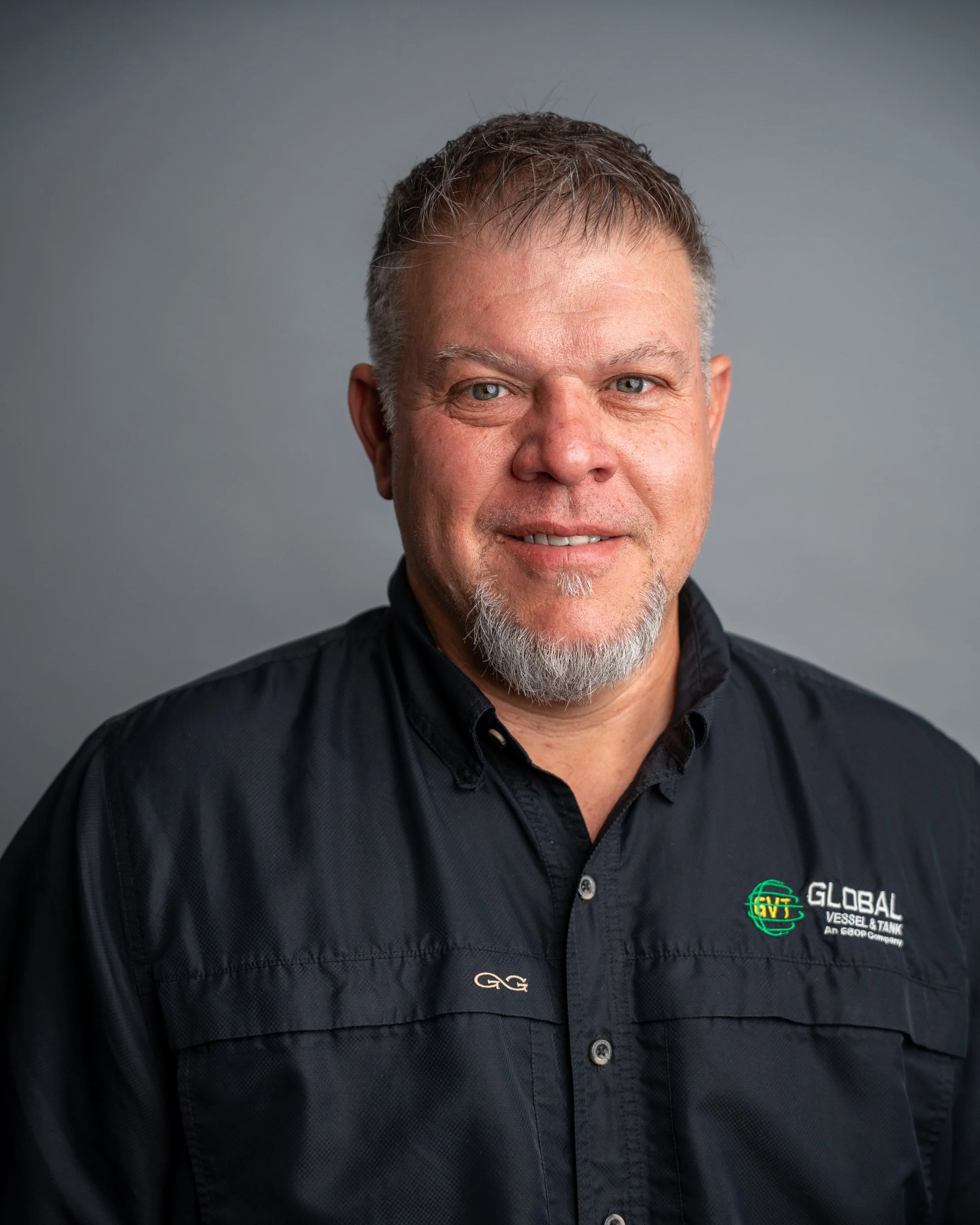 Close-up of a middle-aged man's face with gray hair and beard, wearing a black button-up shirt with logos and text on the chest, smiling slightly against a light gray background.