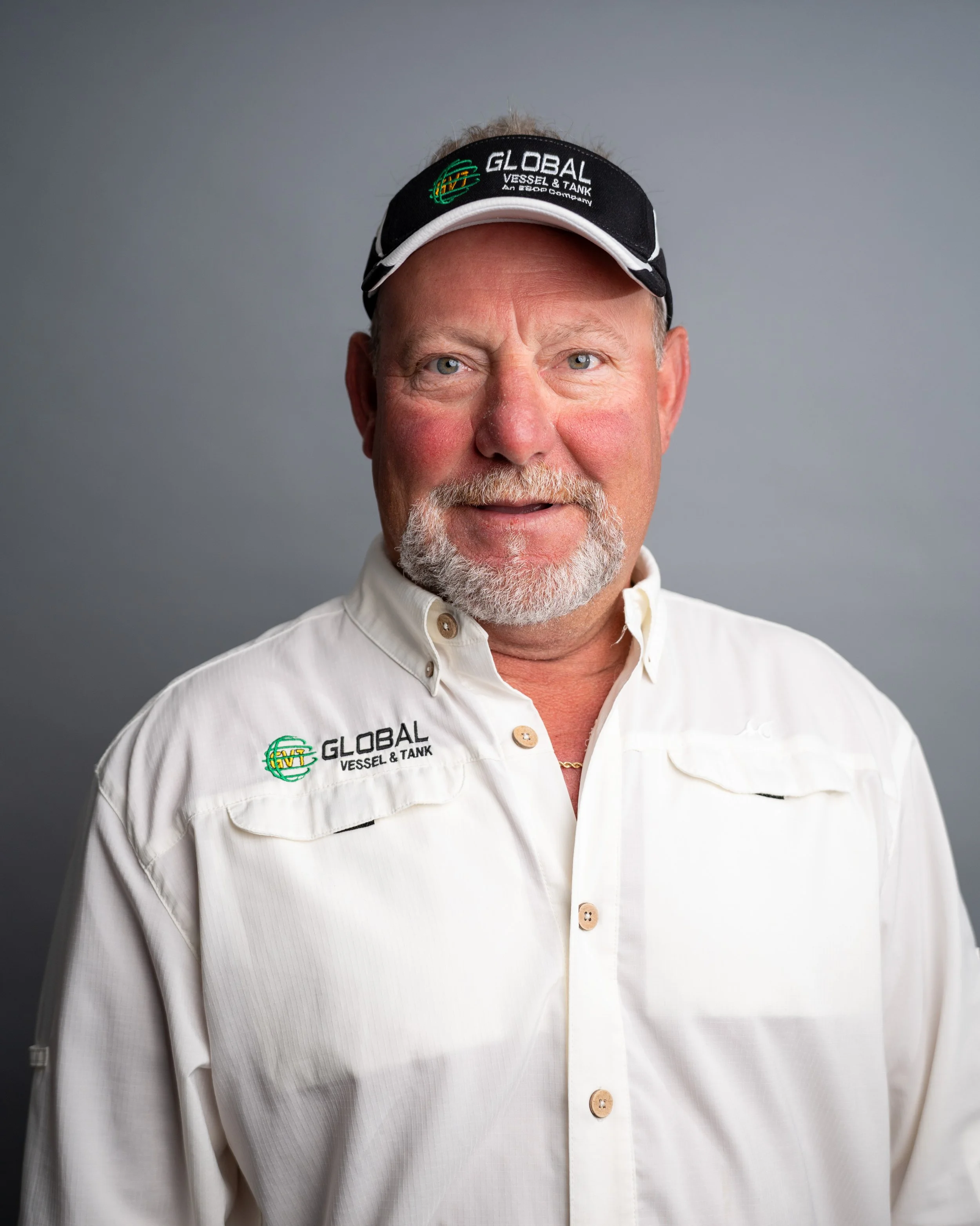A middle-aged man with gray hair and a gray beard wearing a white shirt with logo embroidery and a black cap with a logo and text, standing against a plain gray background.