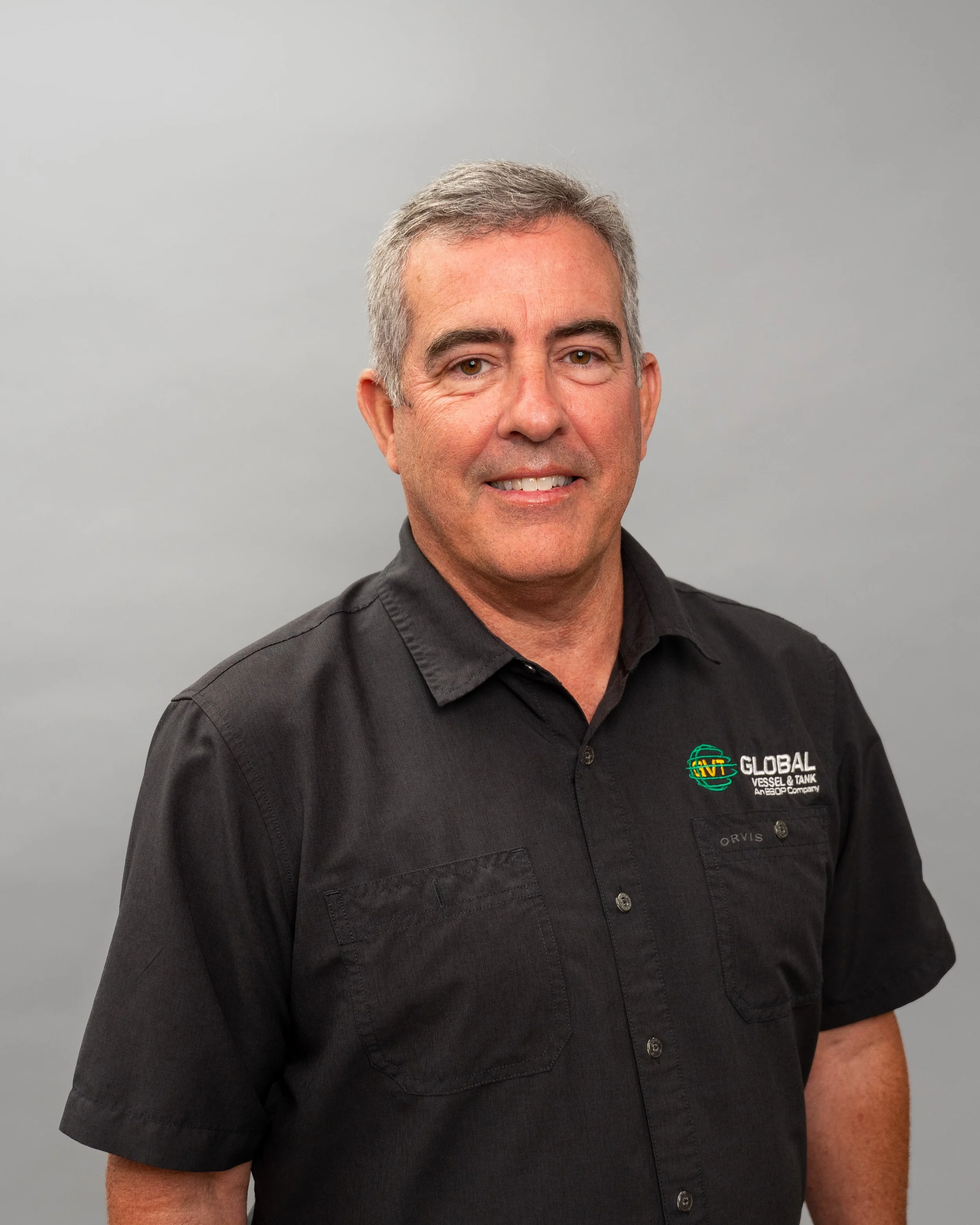 A middle-aged man with short gray hair wearing a black collared shirt with a company logo, standing against a plain gray background.