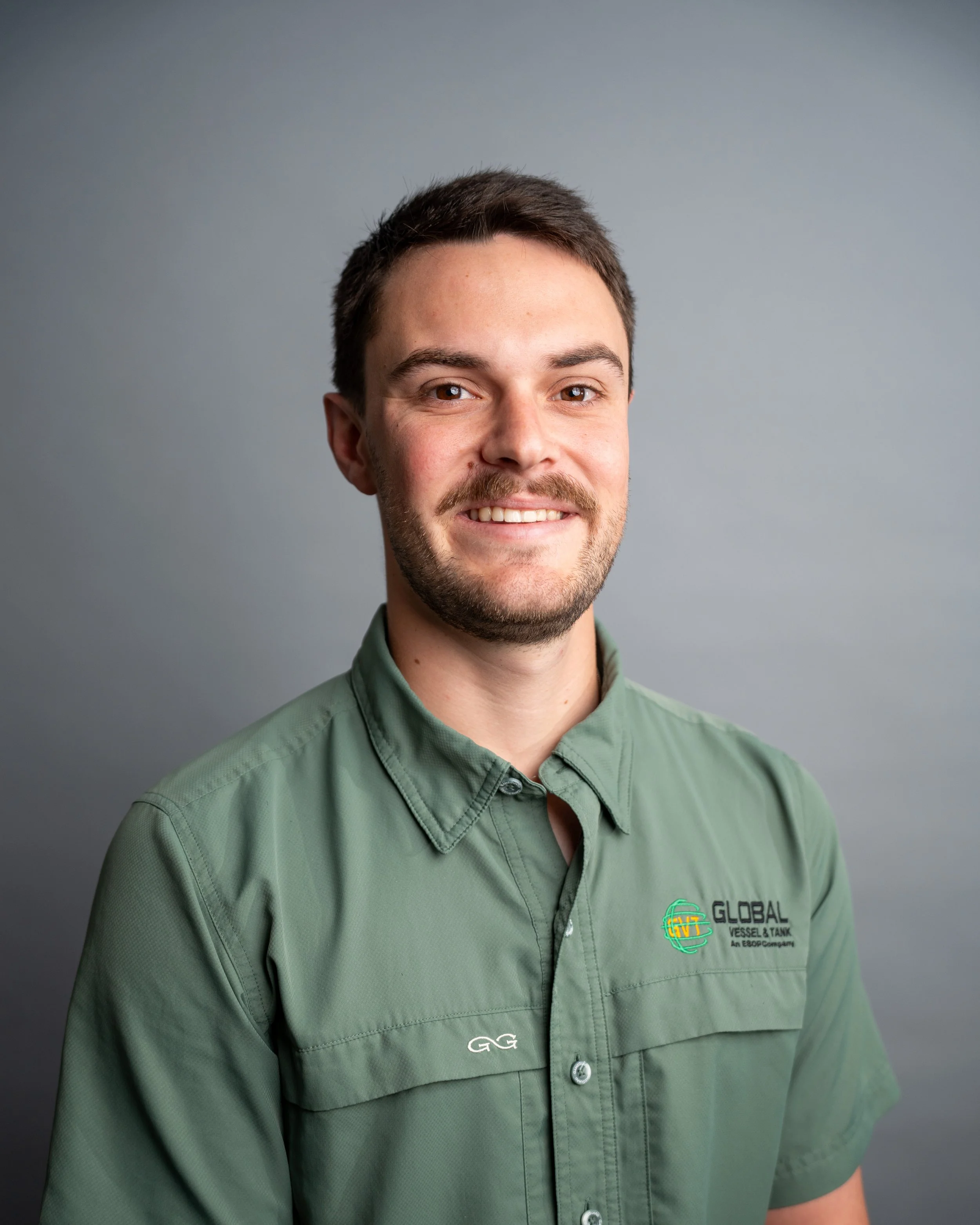 A young man with short dark hair, light skin, and a beard, smiling and wearing a green shirt with a company logo on the chest, in front of a gray background.