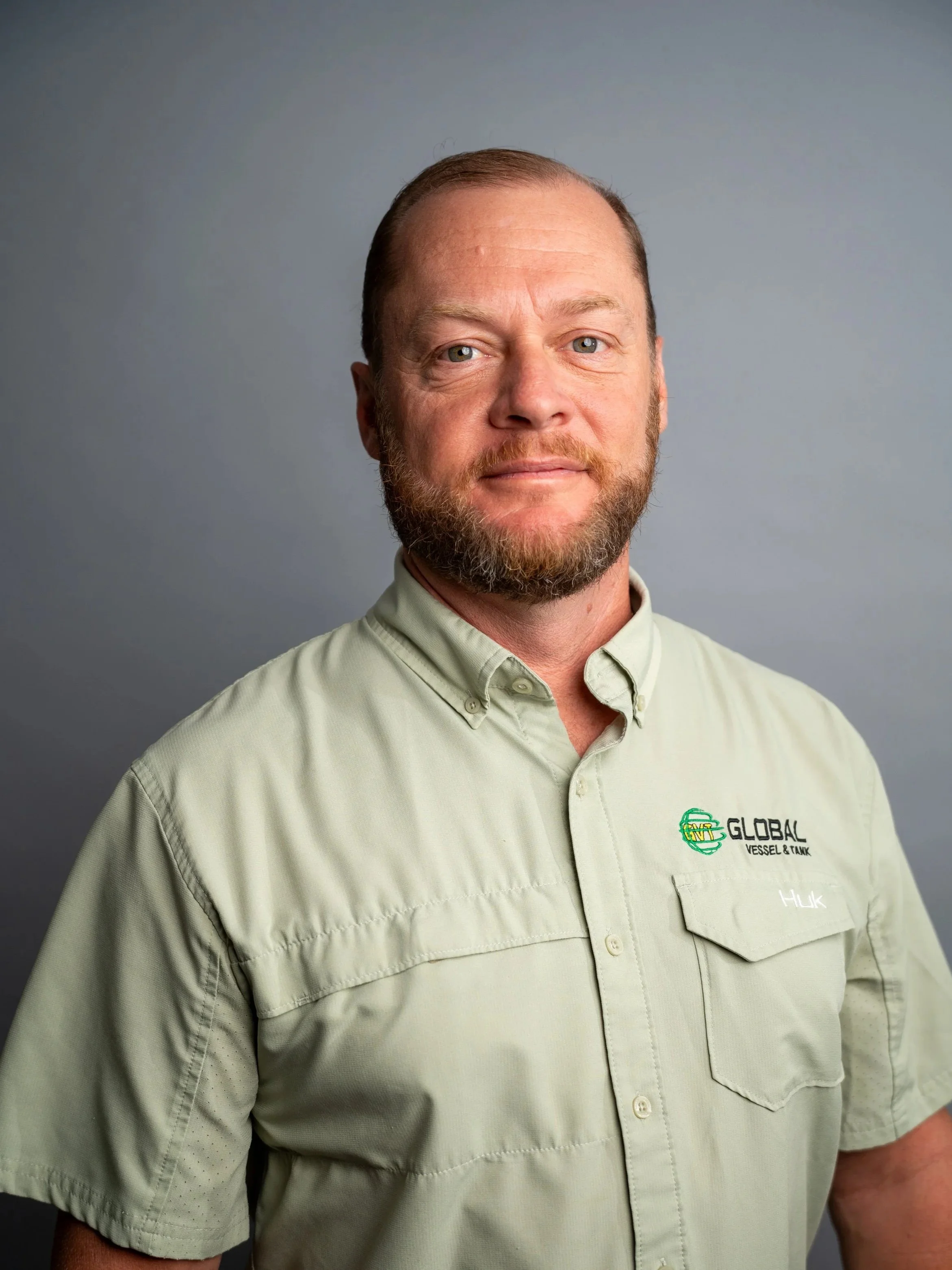 A man with short brown hair, beard, and mustache wearing a light green shirt with a company logo on the chest. The background is plain gray.