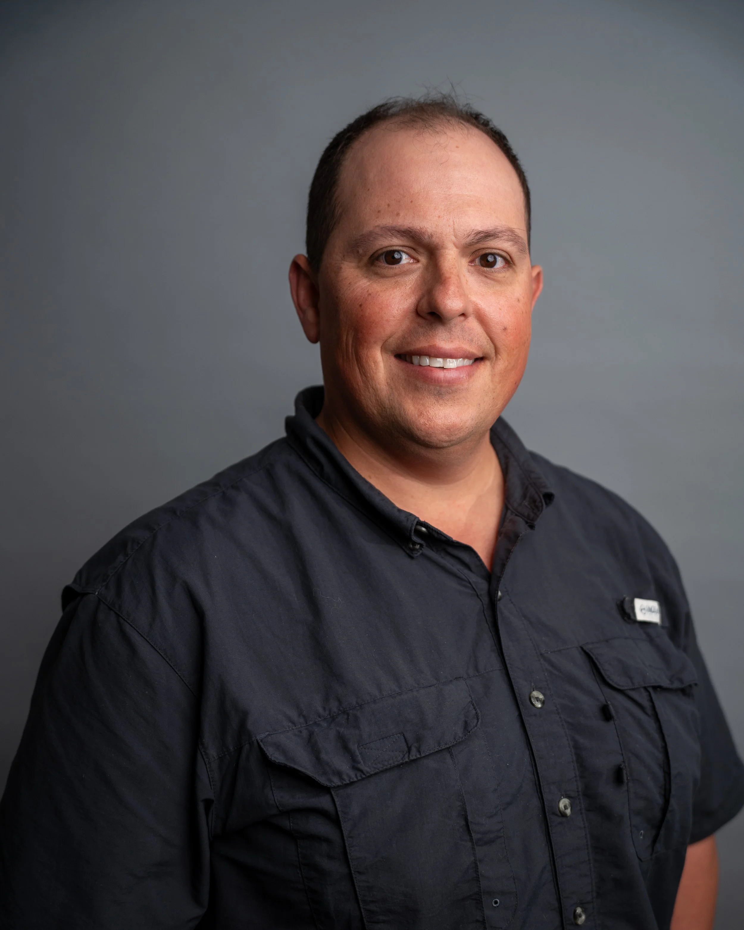Close-up photo of a smiling man with short dark hair, brown eyes, wearing a dark blue button-up shirt, against a plain gray background.