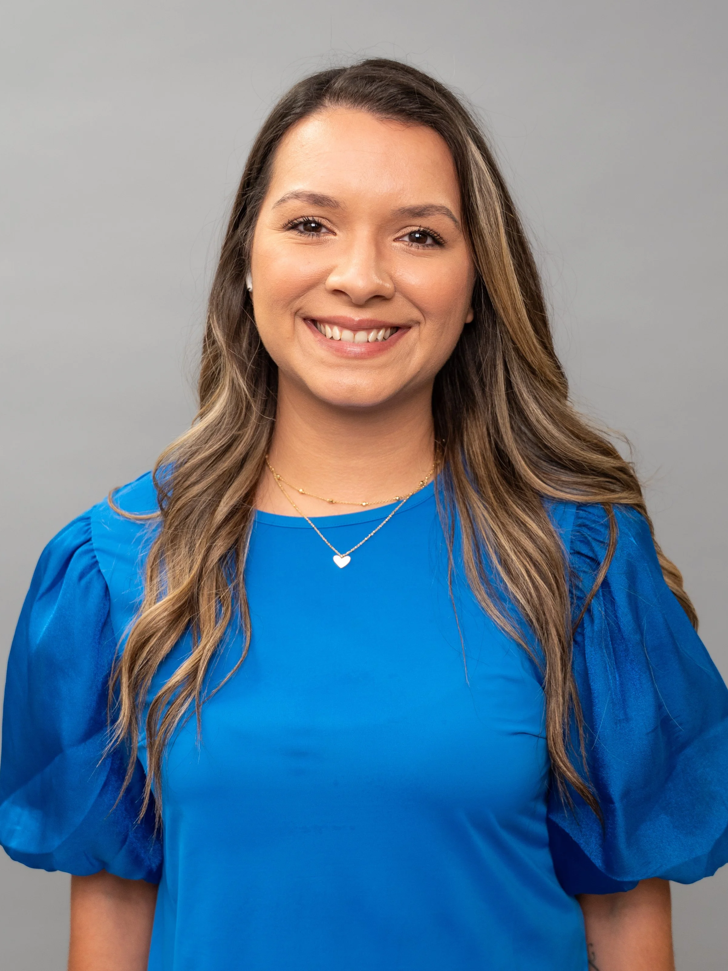 A woman smiling, wearing a blue top with short puffed sleeves, gold jewelry, and a heart-shaped necklace, standing against a gray background.