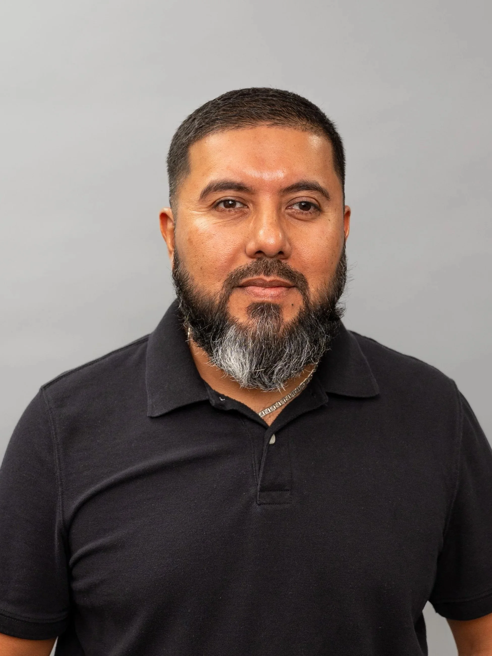 Close-up of a man with a beard and short hair wearing a black polo shirt against a gray background.