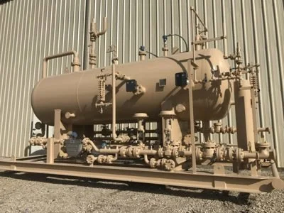 Industrial equipment, a large beige cylindrical tank with pipes and gauges, mounted on a rail track in front of a metal wall.