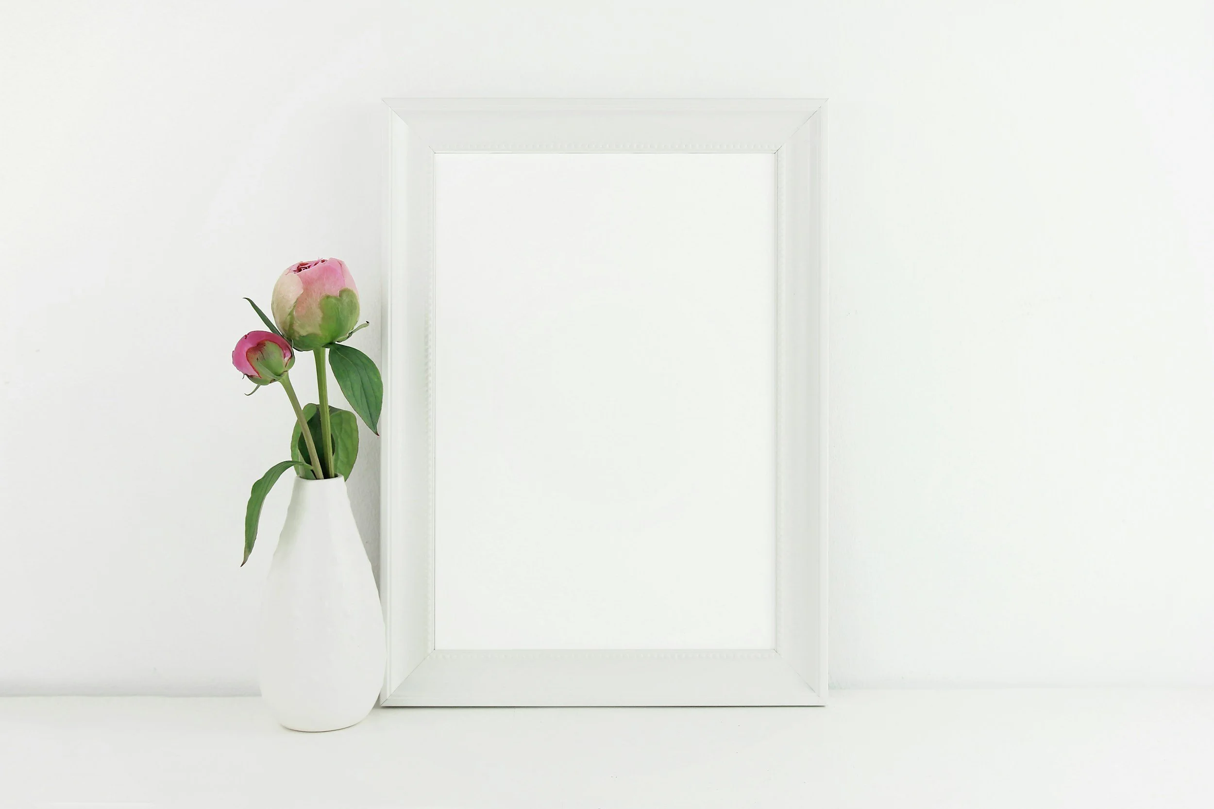 A white vase with pink peonies placed on a white surface next to a white empty picture frame against a plain white background.