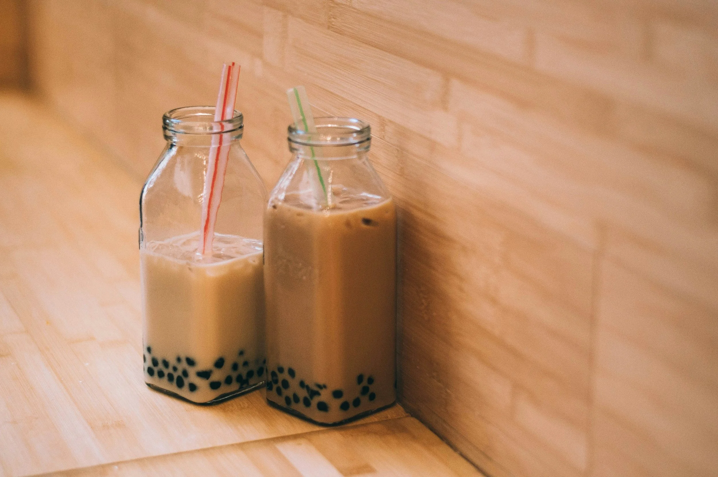 Two glass bottles of bubble tea with colorful striped straws, one with black tapioca pearls at the bottom, placed on a wooden surface against a wooden wall.