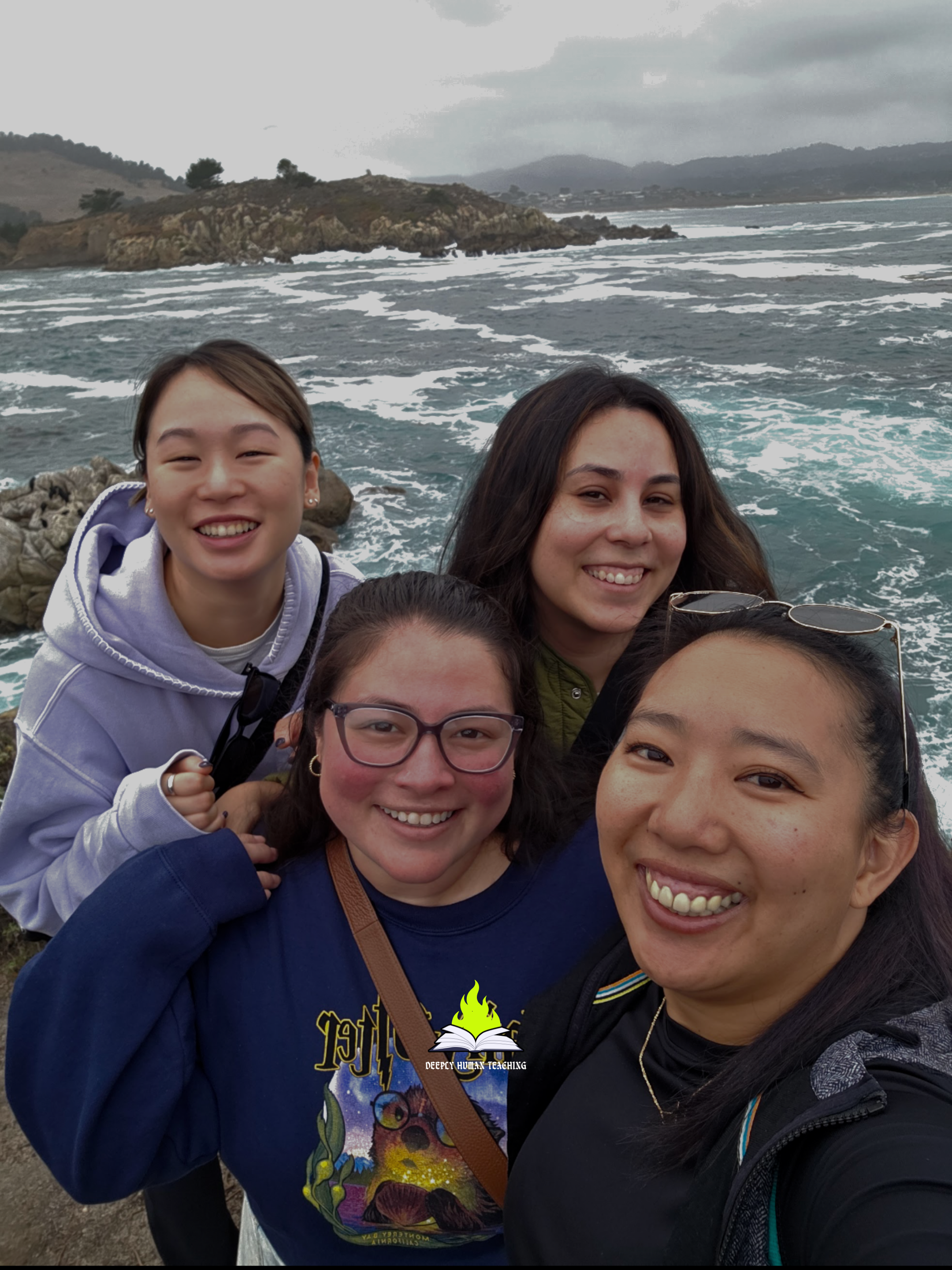 Group of five people taking a selfie on a rocky coastline with ocean waves and an island in the background on a cloudy day.