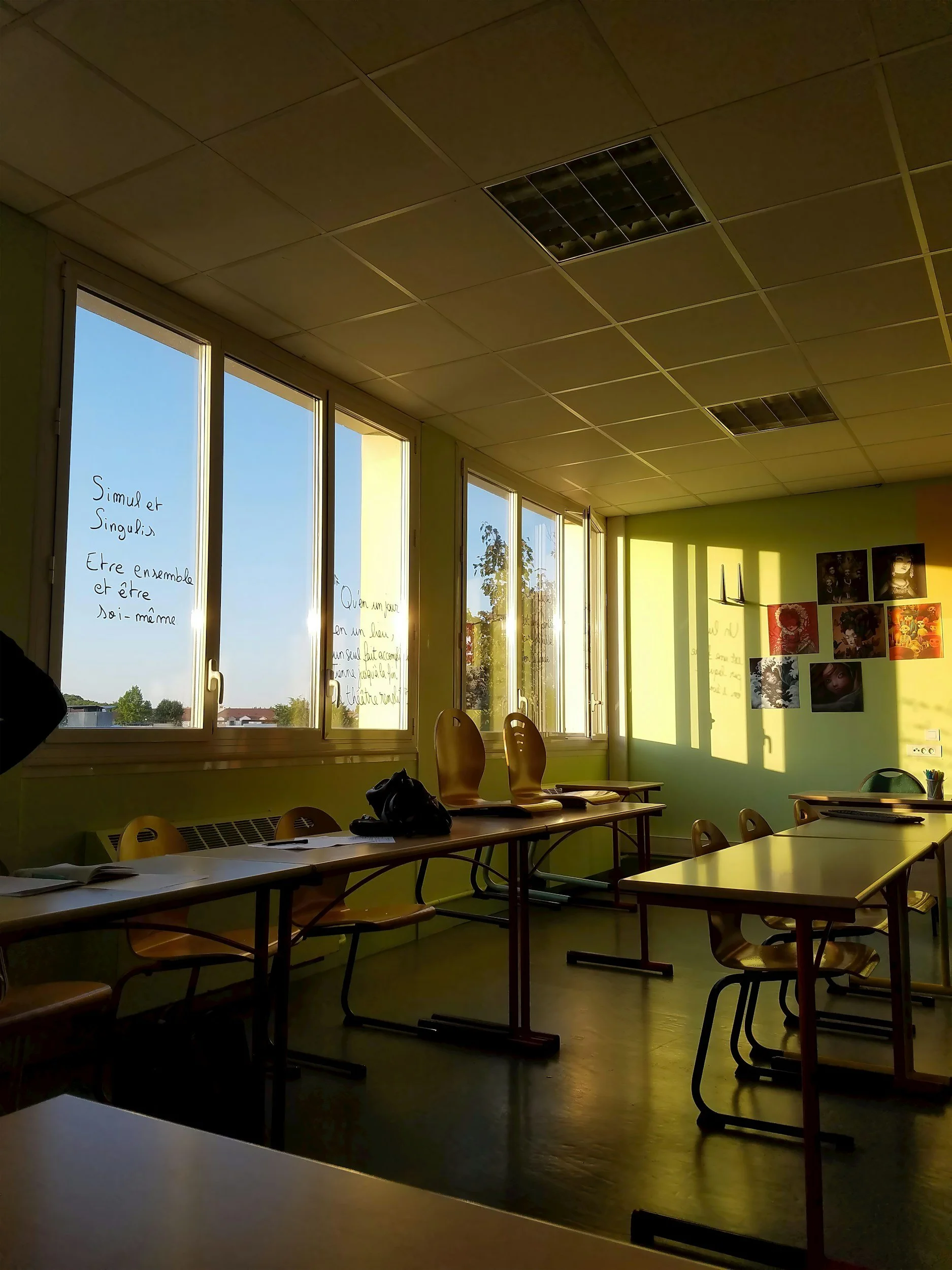 Empty classroom with sunlight streaming through large windows, light green and yellow walls adorned with photographs and writings, and light wood tables and chairs.