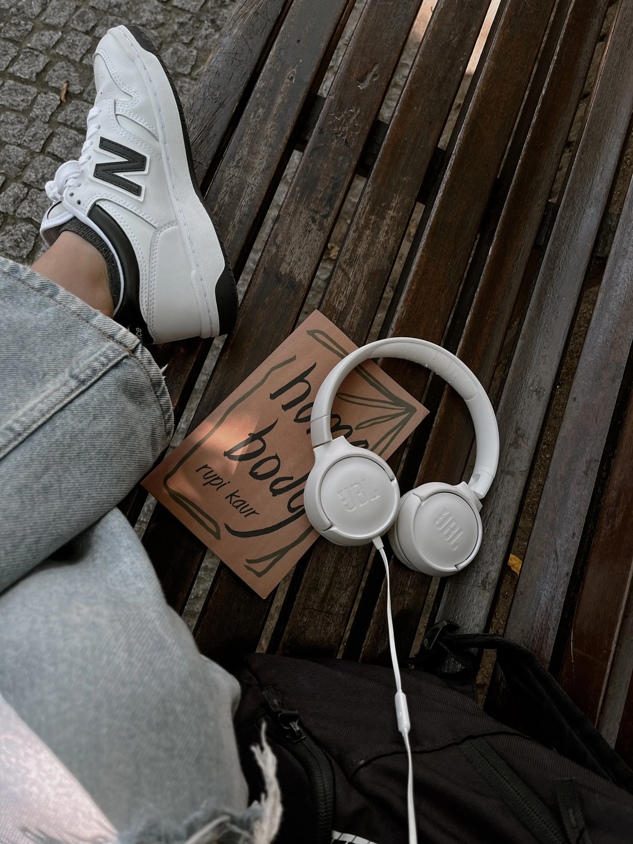 Person sitting on a wooden bench wearing gray ripped jeans, black sneakers, and white socks, with white JBL headphones and a brown shopaholic paper bag on the bench.