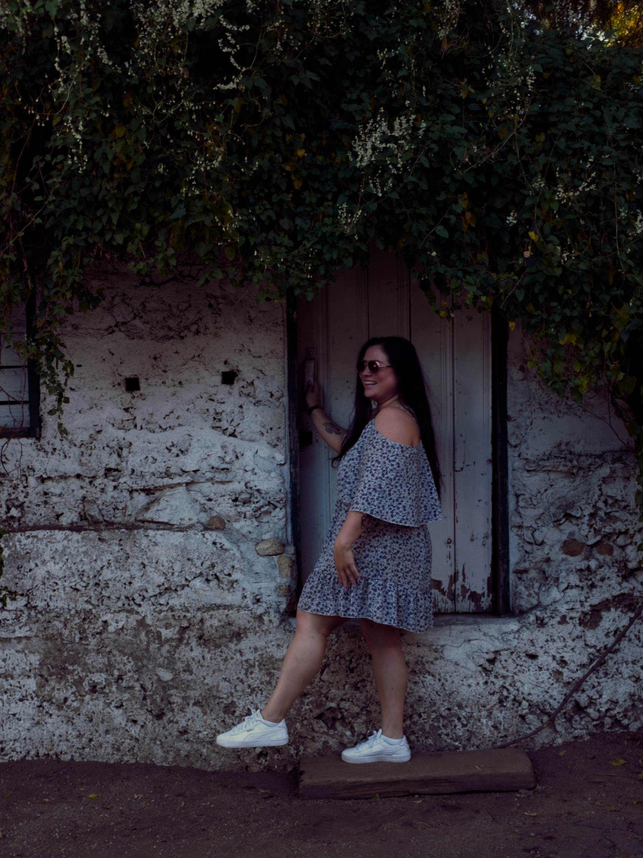 A woman with long dark hair and sunglasses is smiling, wearing a floral dress and white sneakers, standing in front of a weathered wall with a wooden door and green foliage overhead.