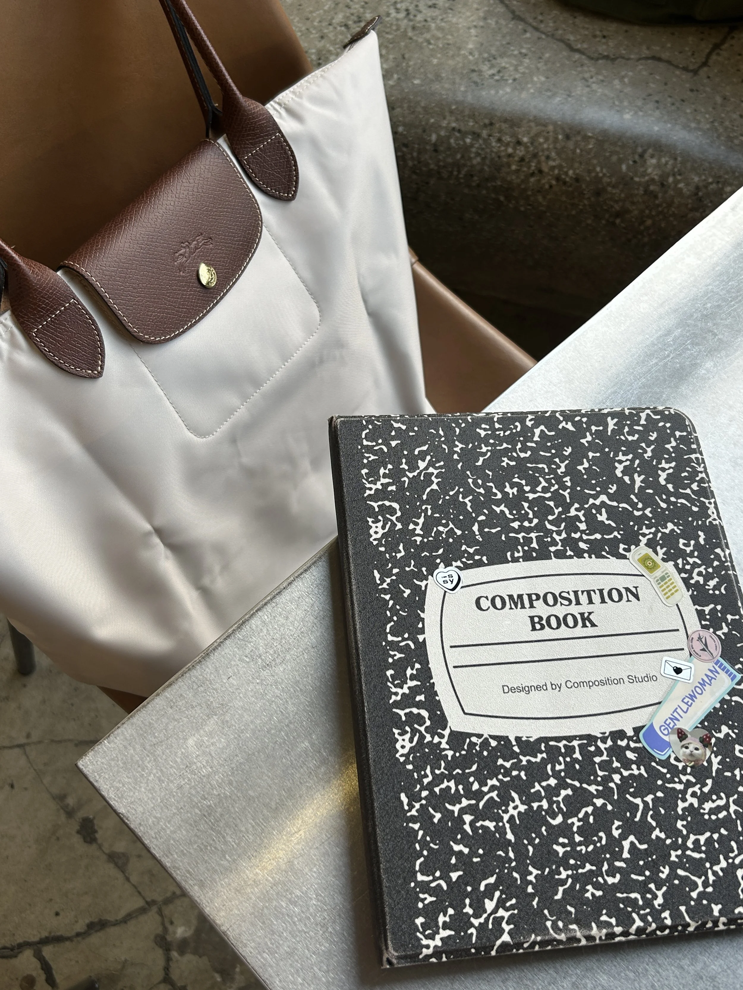 A white tote bag with brown leather accents and a black and white composition notebook on a metal table. There are also several colorful pins attached to the notebook, including a dog, a yellow flag, and a woman, with some text visible.