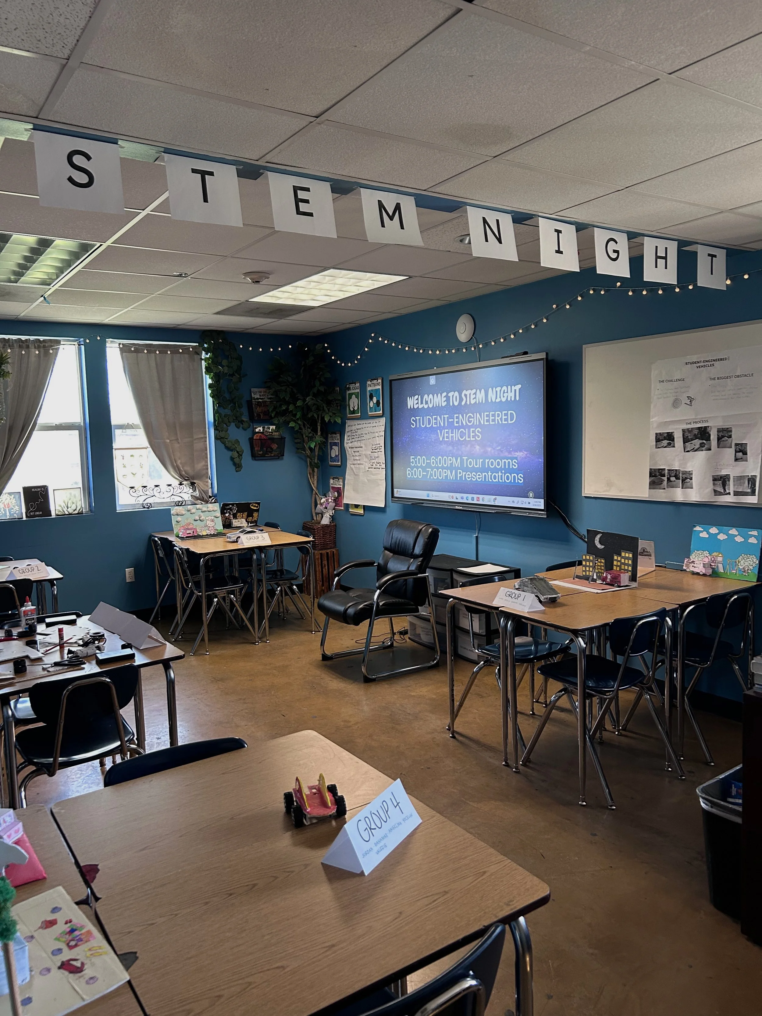 A classroom set up for a STEM night event with tables labeled for different groups, a large screen displaying event details, and decorations including string lights and posters.