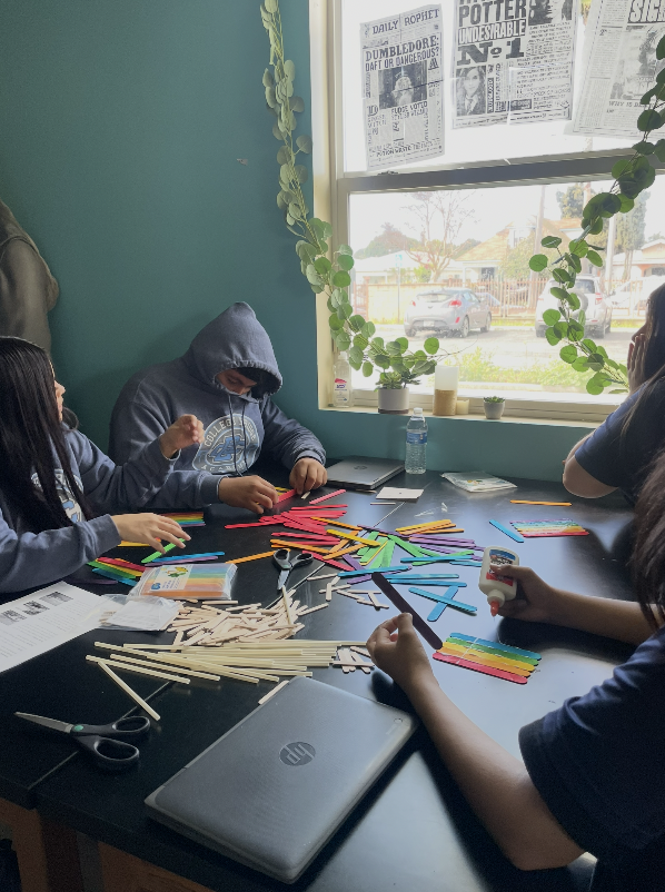 Group of children working on arts and crafts project with colorful popsicle sticks at a table inside a room with a teal wall, large window, and newspaper posters.