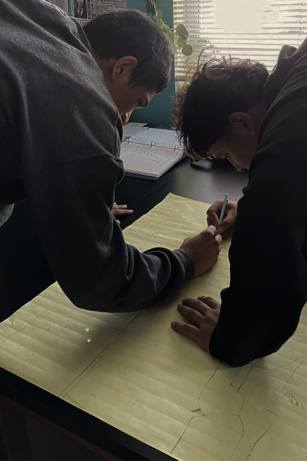 Two students are working together on a project or poster, writing or drawing on a large sheet of yellow paper. They are bent over the table in a classroom setting with books and a window with blinds in the background.