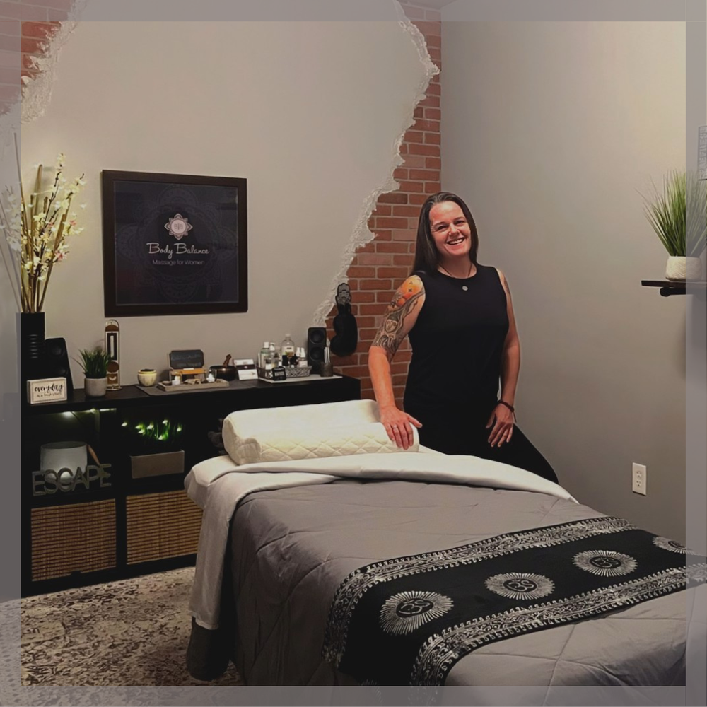 A woman standing beside a massage bed in a wellness or massage therapy room, with decorative plants, a flat-screen TV on the wall, and aesthetic decor.