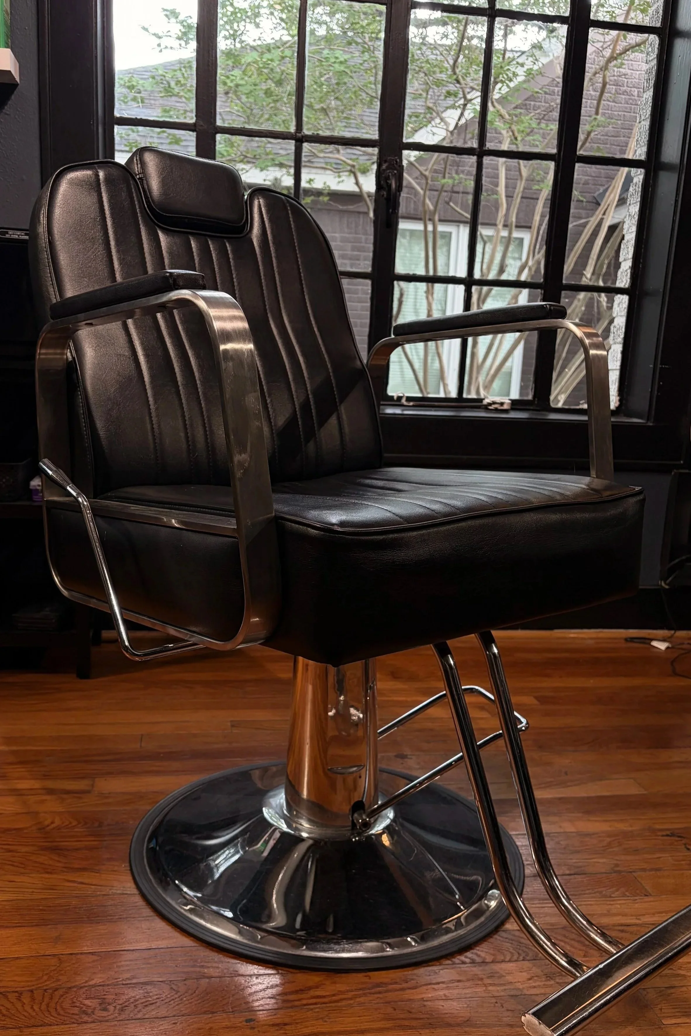 A black leather barber chair with metal armrests and a chrome base in front of a large window with black frames, wooden flooring inside a barbershop or salon.